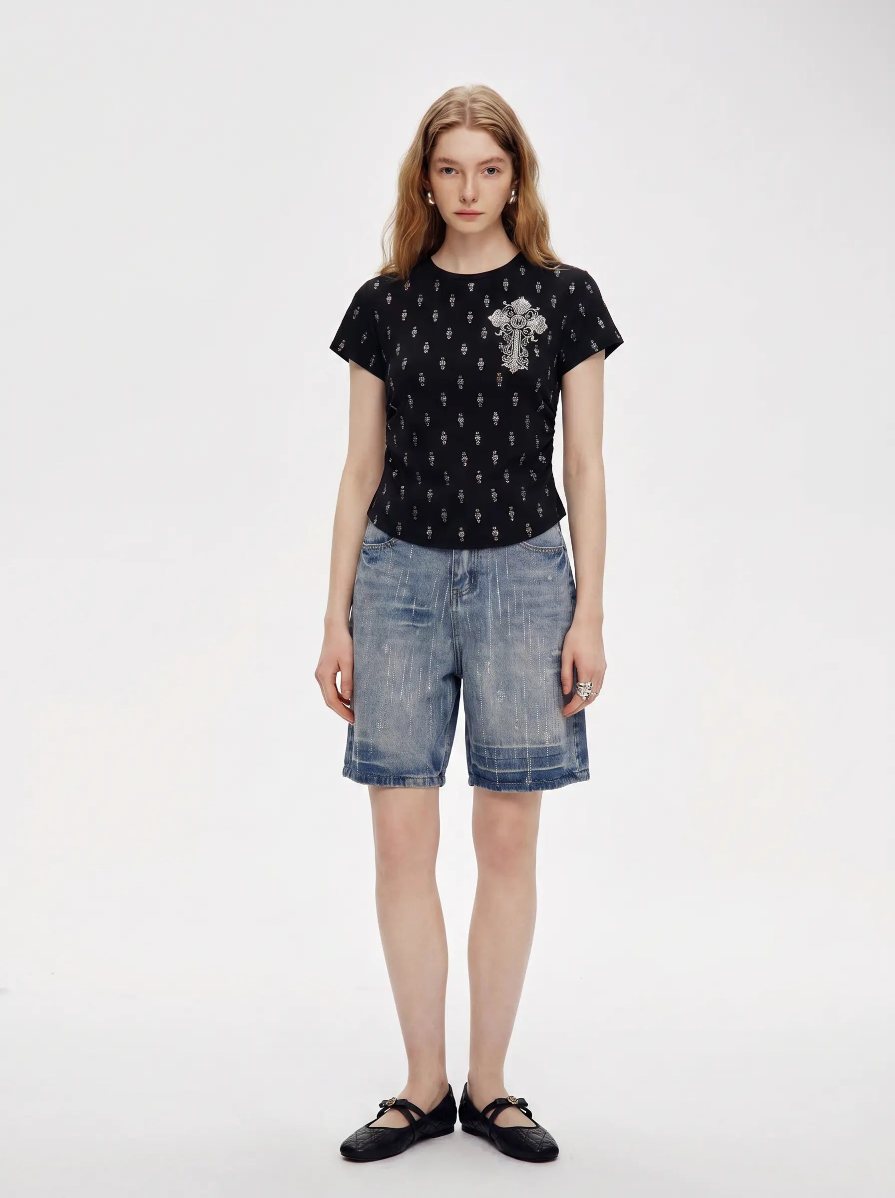 A model stands against a plain light gray studio background wearing a black short-sleeve t-shirt with a small white graphic on the chest, blue denim shorts, and black flat shoes. TCH Rhinestone Techno Cross Graphic Black T-Shirt