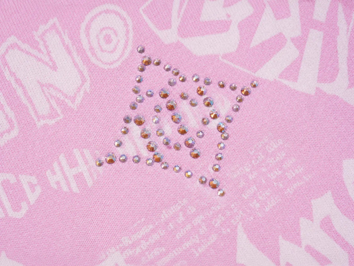 A close-up of a pink fabric surface with a decorative arrangement of small iridescent rhinestones forming a triangular shape over faint white graphic text. TCH Rhinestone Techno Graphic Wide-Leg Sweatpants