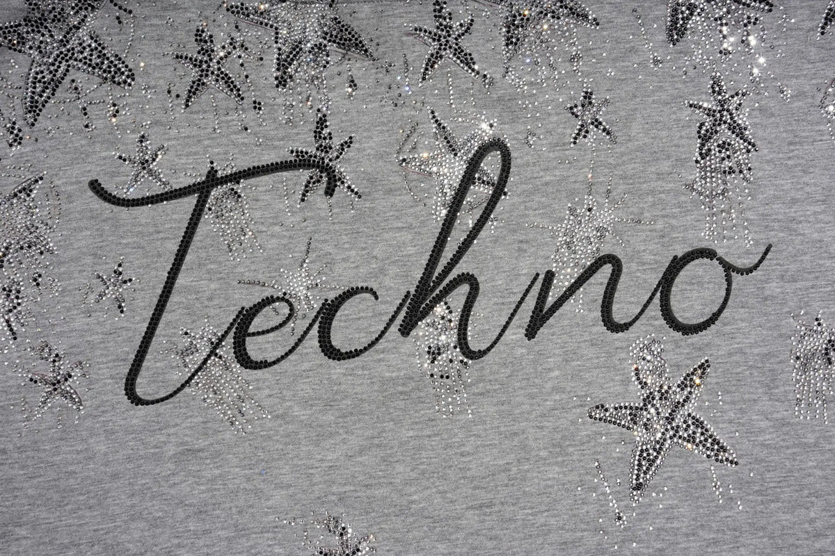 Close-up of a gray fabric surface with the word "Techno" in black script and scattered silver rhinestone star embellishments. TCH Rhinestone Techno Unicorn Graphic T-Shirt