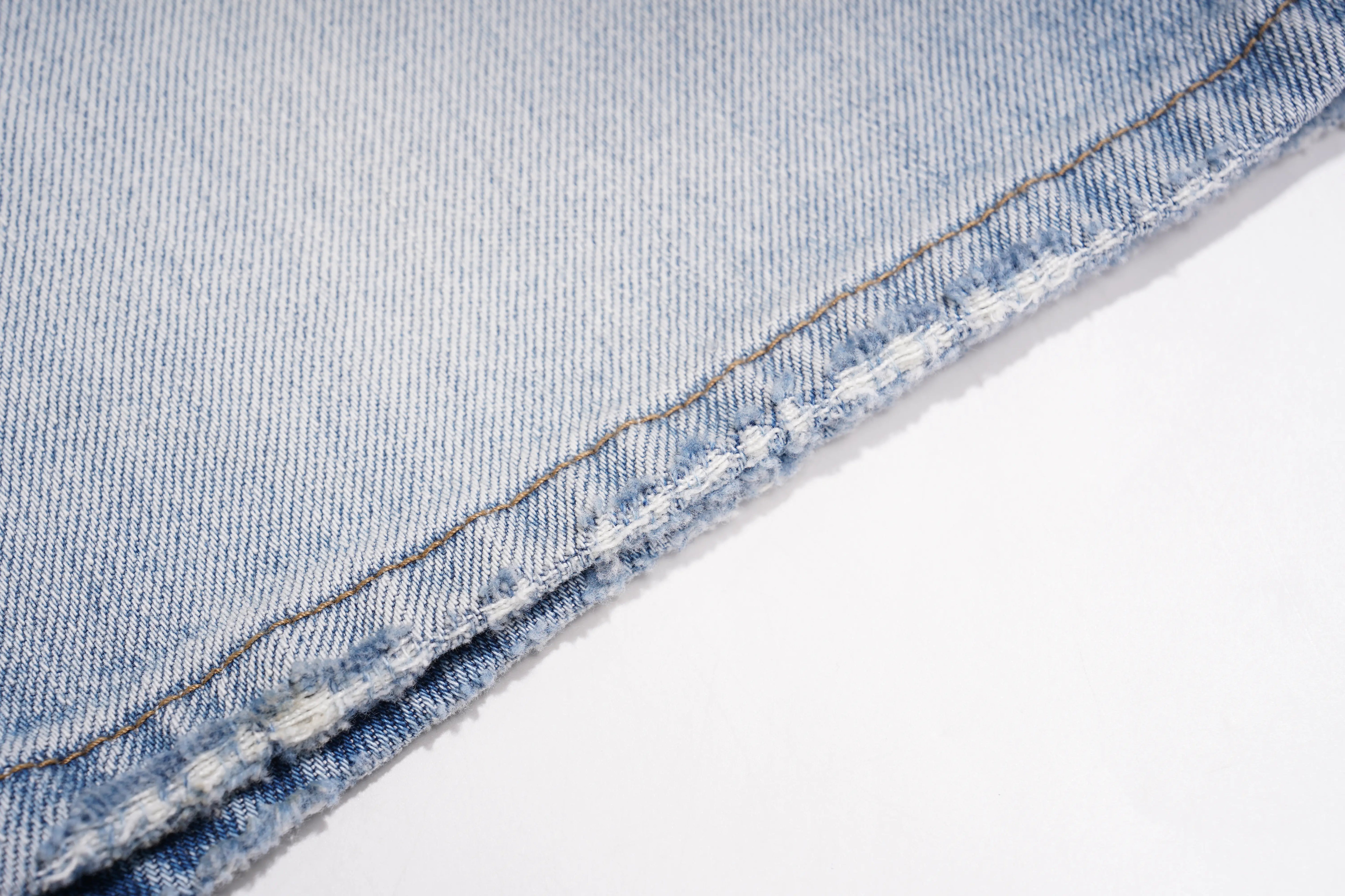 Close-up of a light blue denim fabric corner showing the woven texture and double-stitched raw hem on a white background. TCH Rhinestone Teddy Bear Bow Denim Shorts