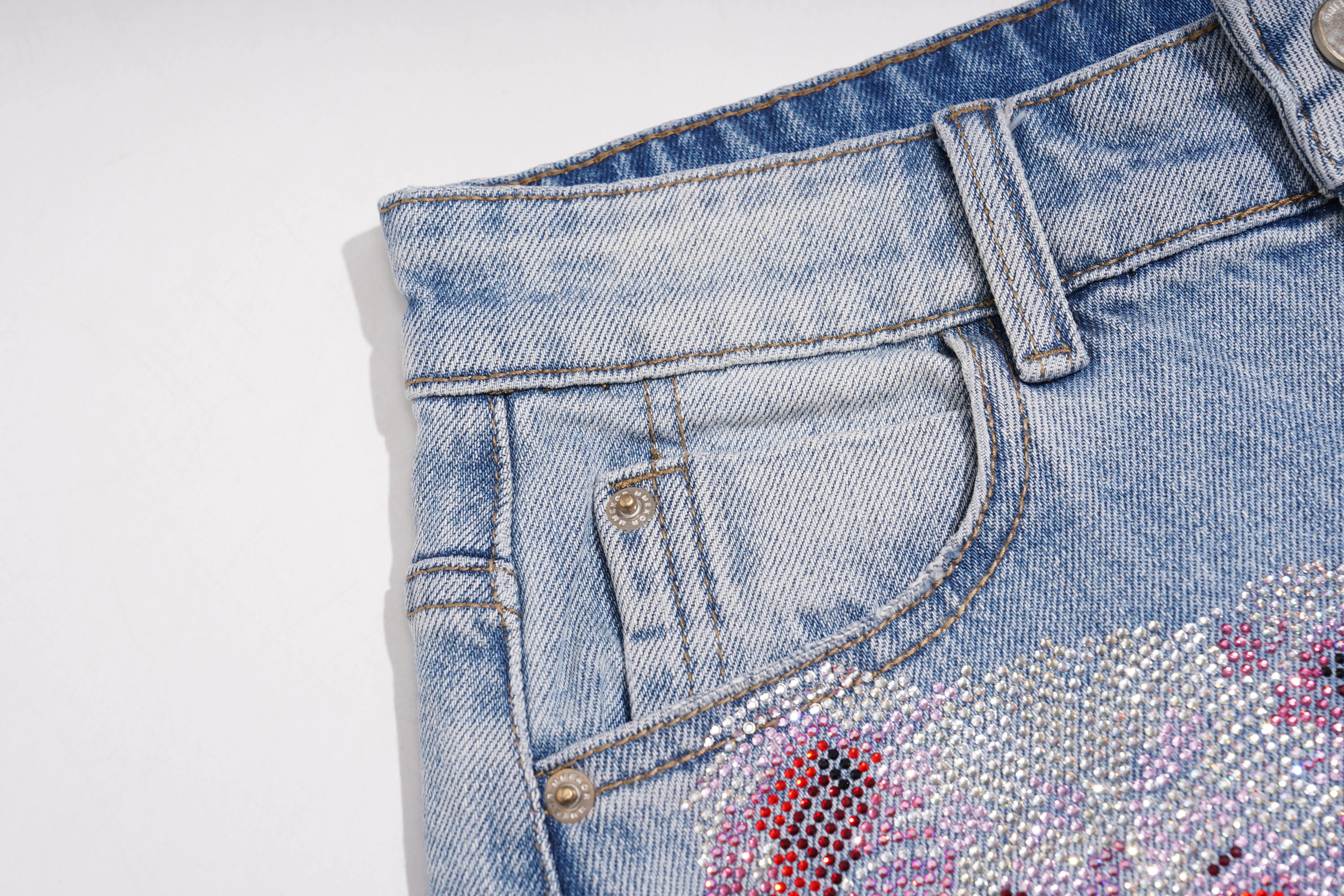 Close-up of the back waistband and pocket area of light blue denim jeans, showing a decorative floral embellishment covered in pink, purple, and silver rhinestones. TCH Rhinestone Teddy Bear Bow Denim Shorts