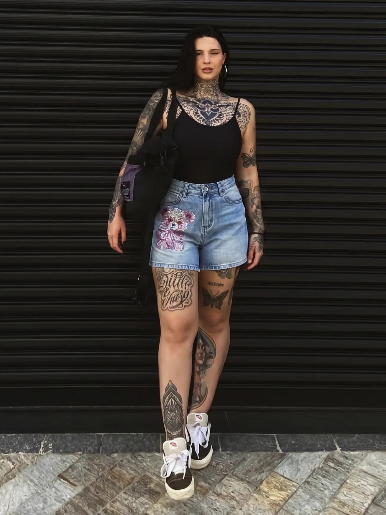 A tattooed woman stands in front of a black metal shutter wearing a black tank top, light-wash denim shorts with floral embroidery, and black-and-white high-top sneakers, while carrying a black shoulder bag. TCH Rhinestone Teddy Bear Bow Denim Shorts