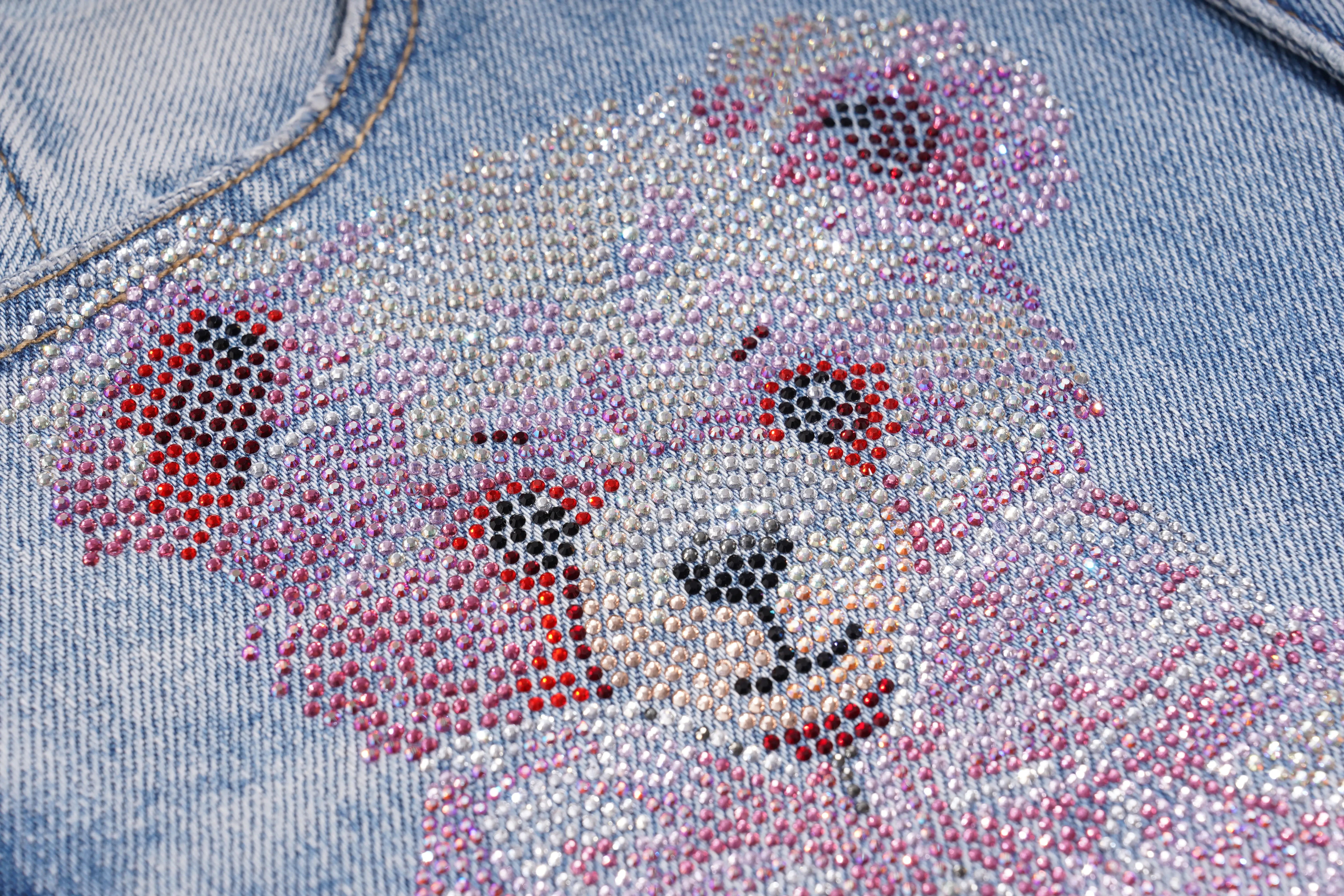 Close-up of a light blue denim garment featuring a teddy bear face embellished with rhinestones in silver, pink, red, and black. TCH Rhinestone Teddy Bear Bow Denim Shorts