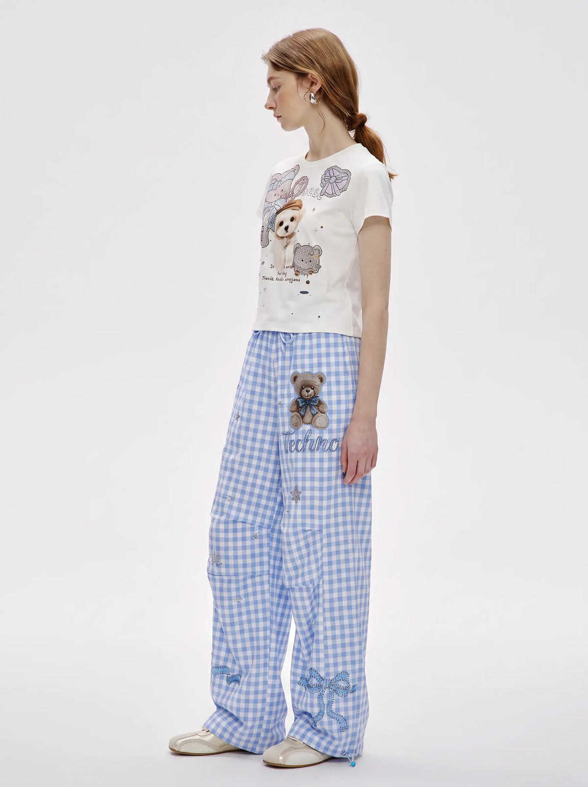 A woman stands in profile wearing a white graphic T-shirt with teddy bear illustrations and light blue-and-white gingham wide-leg pants with a teddy bear patch and bow details near the cuffs. TCH Rhinestone Teddy Bear Bow Gingham Wide-Leg Pants