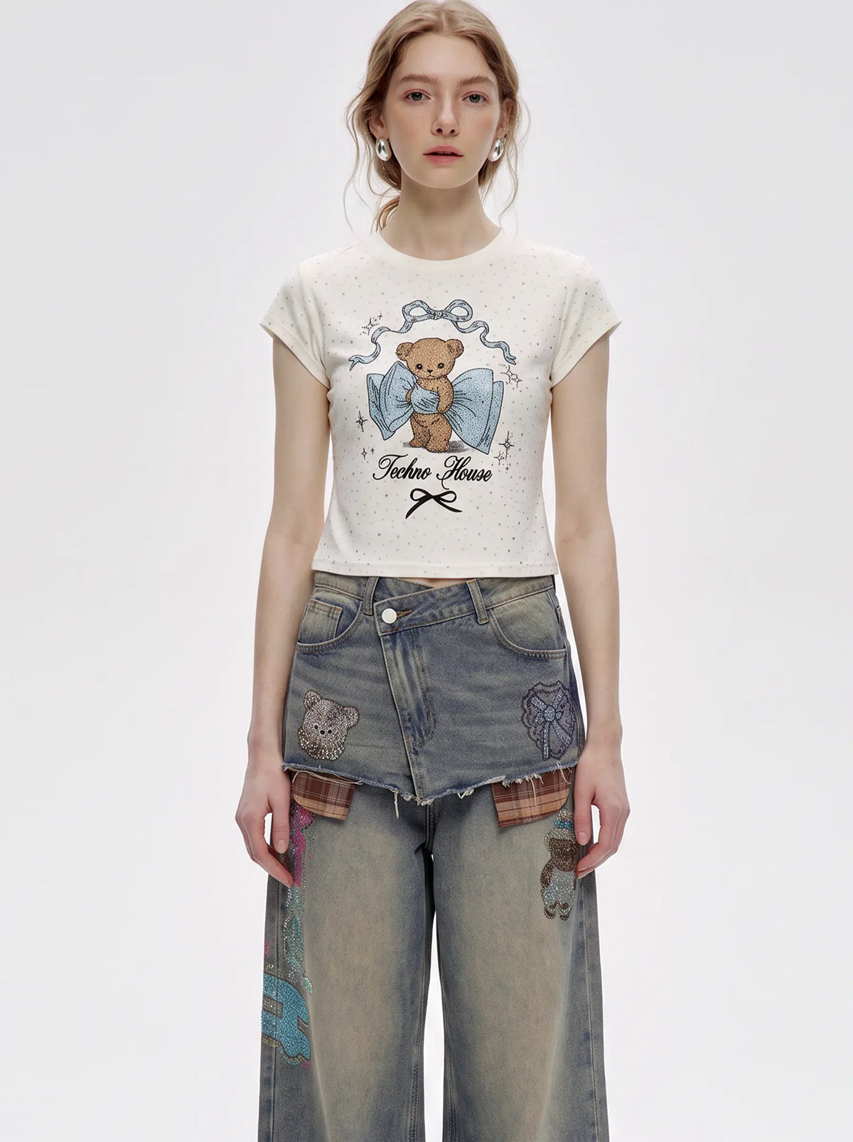 A model wearing a short-sleeve cream-colored graphic t-shirt with a teddy bear illustration and the text 'Fiction House', paired with distressed denim jeans, against a plain light gray studio background. TCH Rhinestone Teddy Bear Bow Graphic Cropped T-Shirt