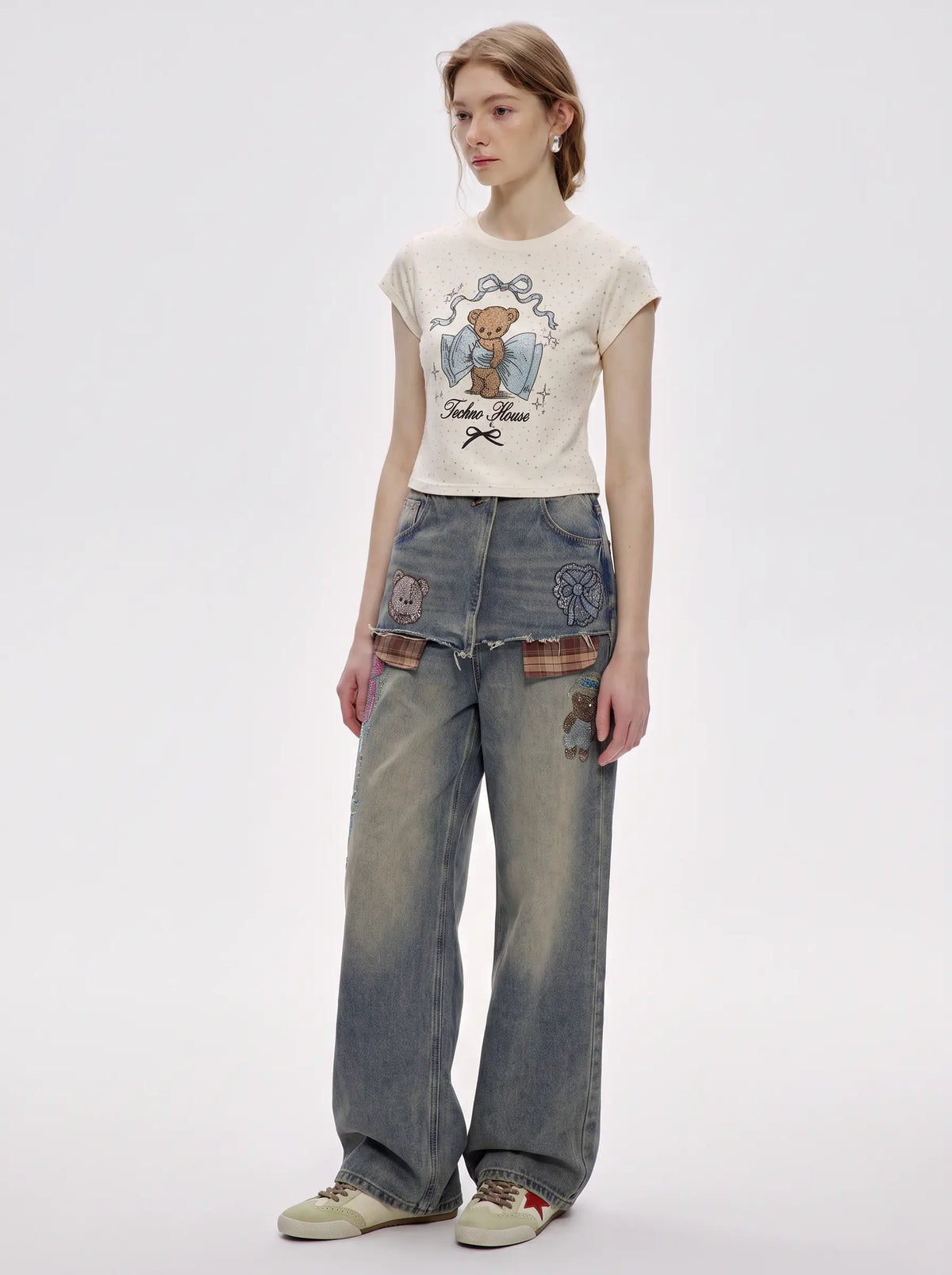 A model stands against a light gray studio background wearing a cream-colored graphic t-shirt with a teddy bear design, wide-leg faded blue jeans with decorative patches, and beige sneakers. TCH Rhinestone Teddy Bear Bow Graphic Cropped T-Shirt