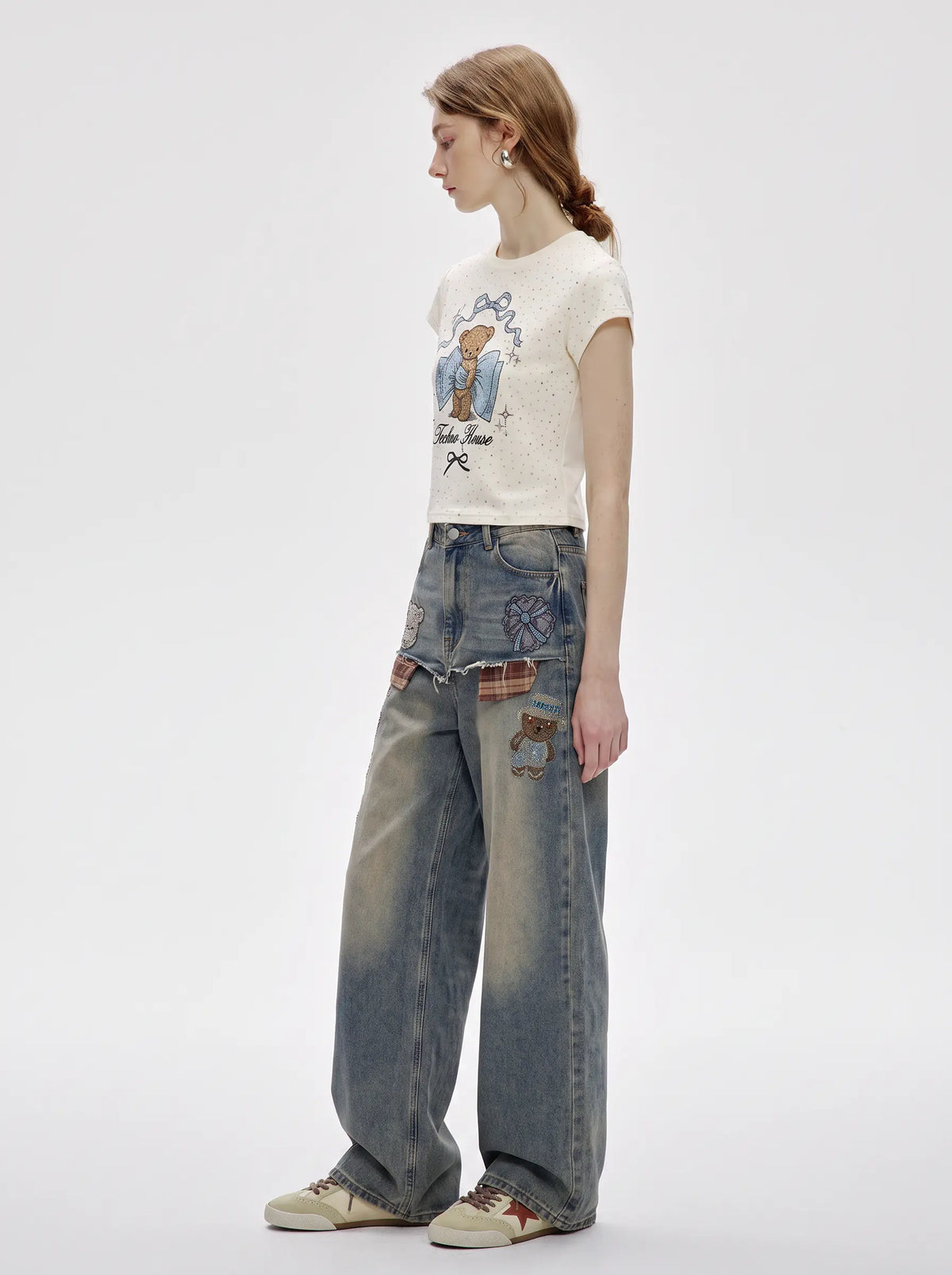 A model is shown from the side wearing a cream-colored short-sleeve t-shirt with a cartoon bear graphic, paired with high-waisted, wide-leg blue jeans featuring patchwork details and beige sneakers, set against a plain light gray studio background. TCH Rhinestone Teddy Bear Bow Graphic Cropped T-Shirt