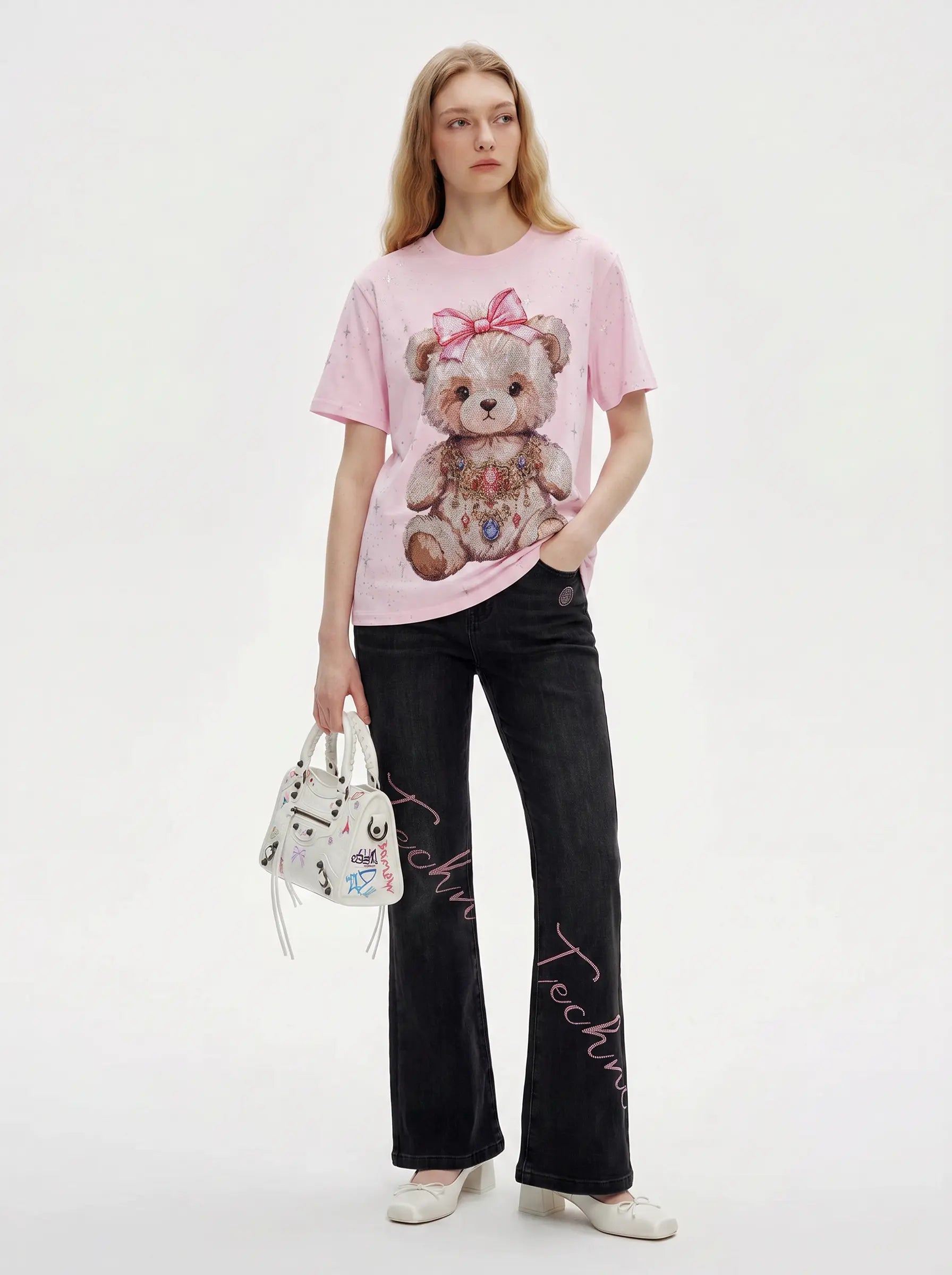 A model stands against a light gray studio background wearing a light pink graphic T-shirt with a teddy bear print, black wide-leg pants with pink script embroidery, white shoes, and holding a small white handbag. TCH Rhinestone Teddy Bear Bow Pink Graphic T-Shirt