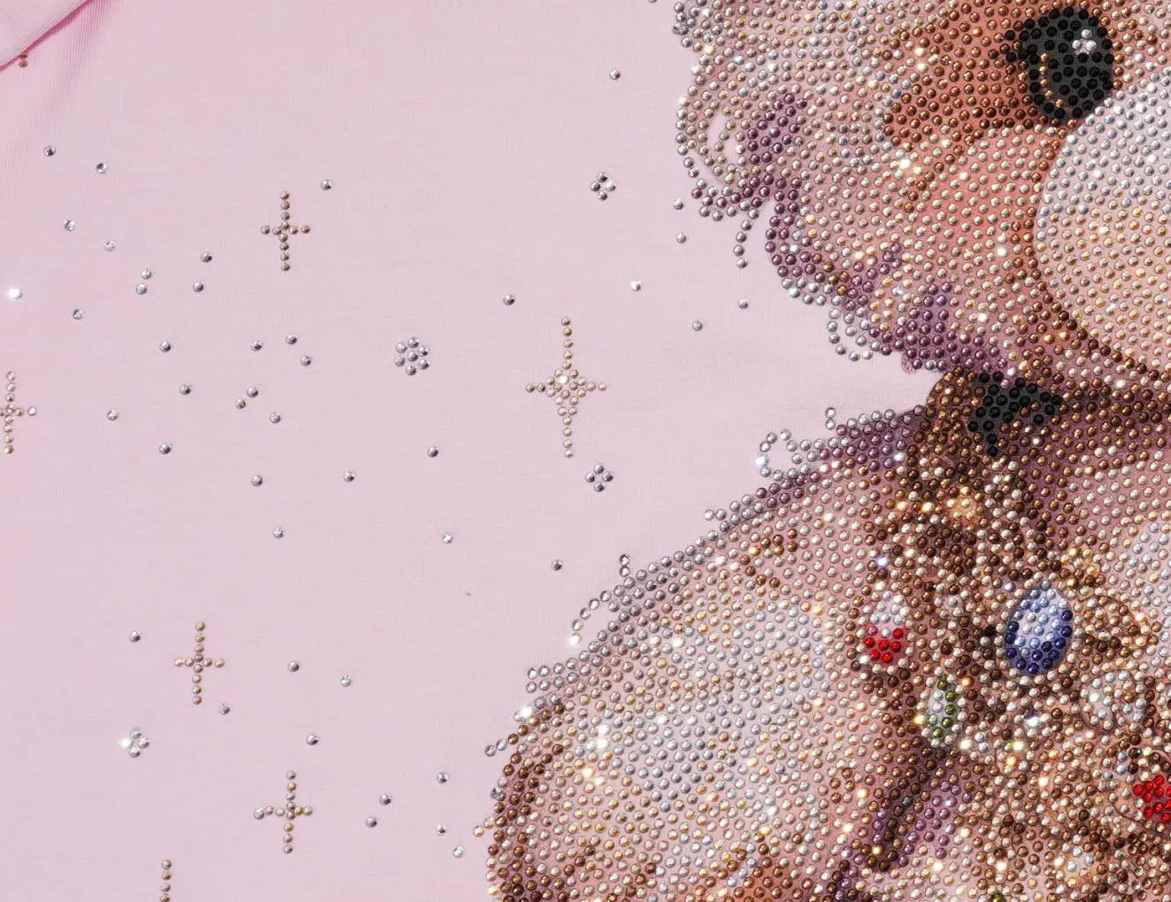 Close-up of a pink surface with a large teddy bear face and upper body made of rhinestones, decorated with jeweled accents, and small rhinestone stars scattered on the background. TCH Rhinestone Teddy Bear Bow Pink Graphic T-Shirt