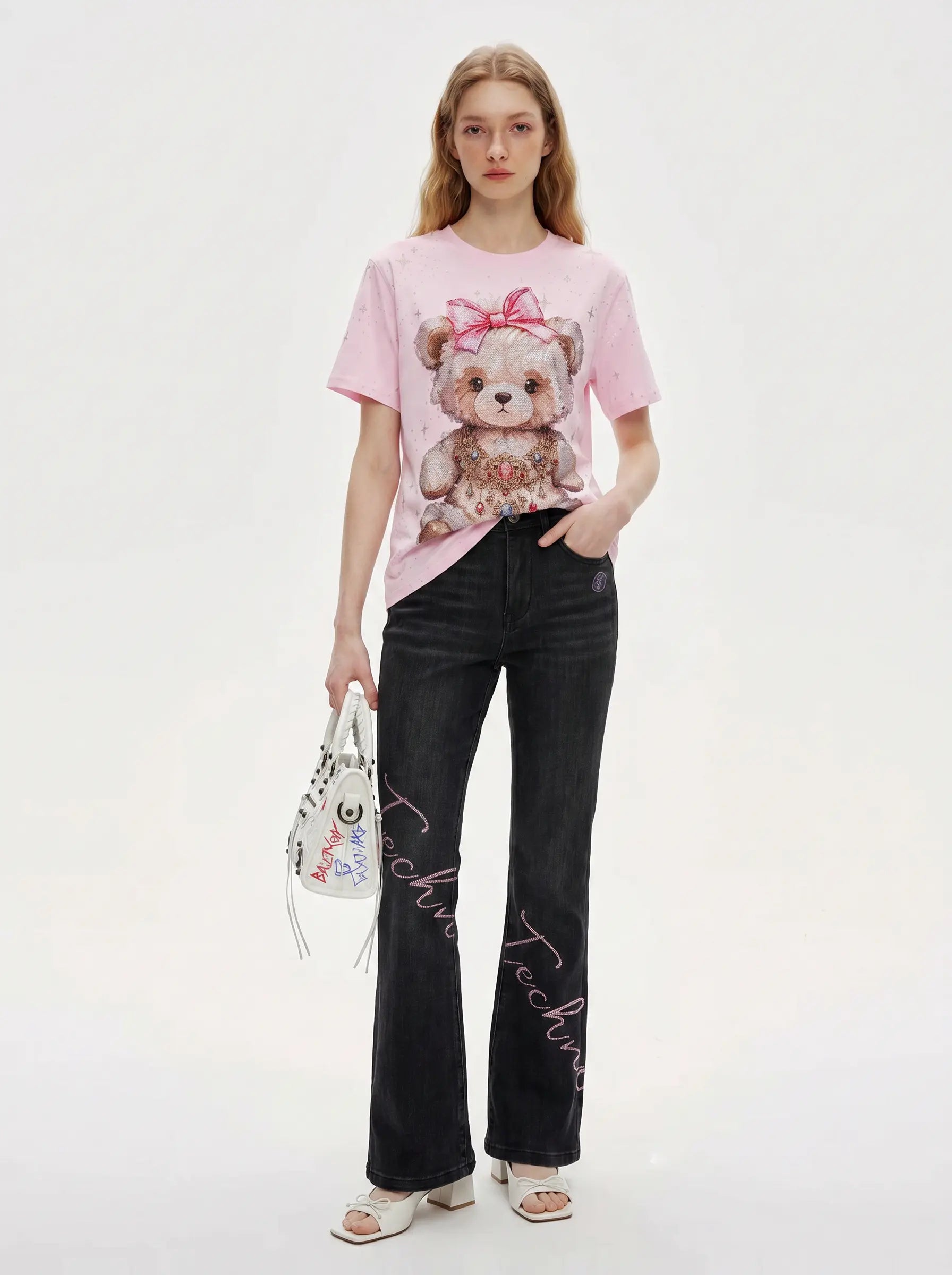 A model stands against a plain white studio background wearing a light pink graphic T-shirt with a teddy bear print, black wide-leg jeans with script embroidery, and white heeled sandals, while holding a small white handbag. TCH Rhinestone Teddy Bear Bow Pink Graphic T-Shirt