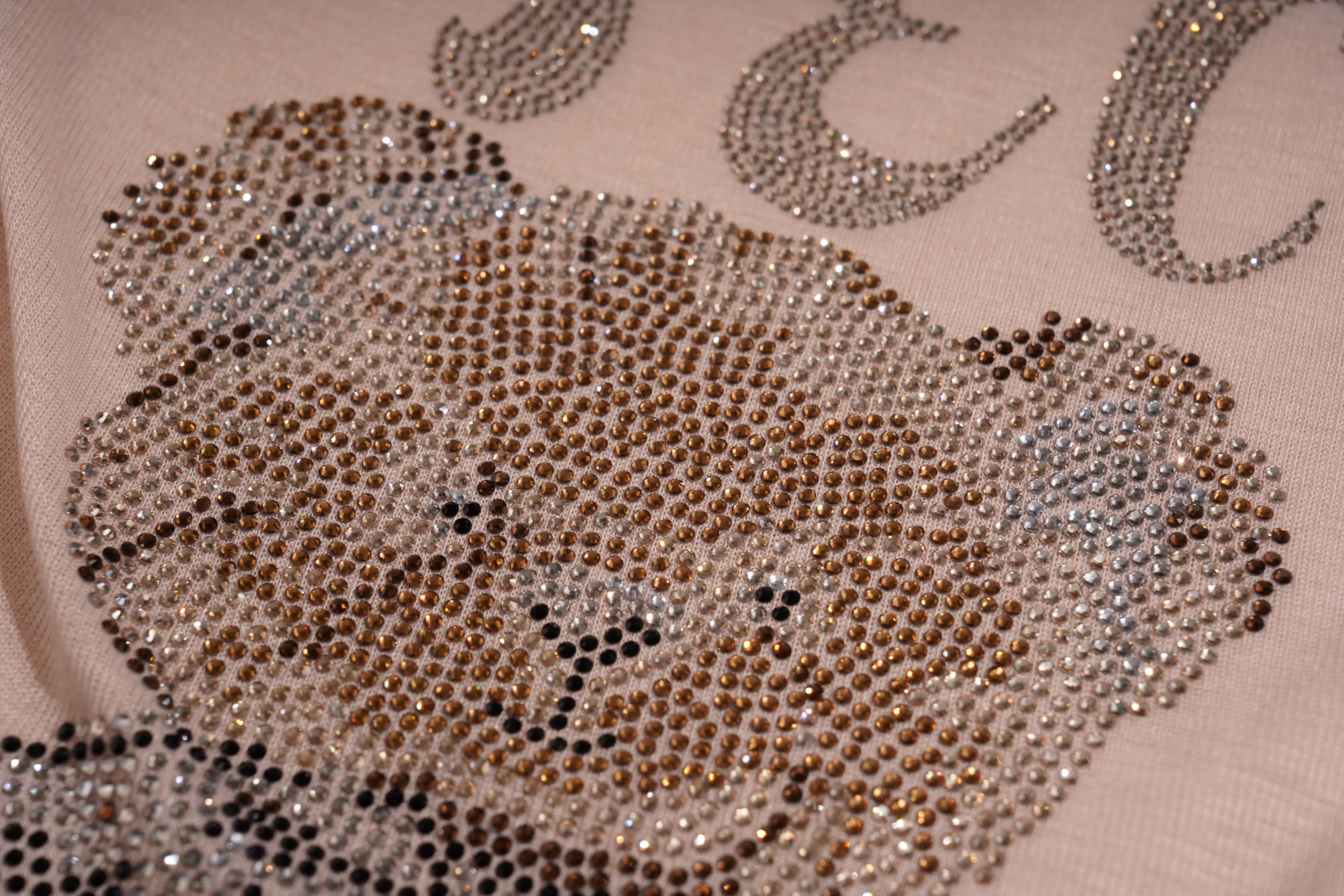 A close-up of a teddy bear face embellished with rhinestones in shades of brown, gold, and silver, on a light beige fabric background with matching lettering. TCH Rhinestone Teddy Bear Bow Ruffle Long Sleeve Top