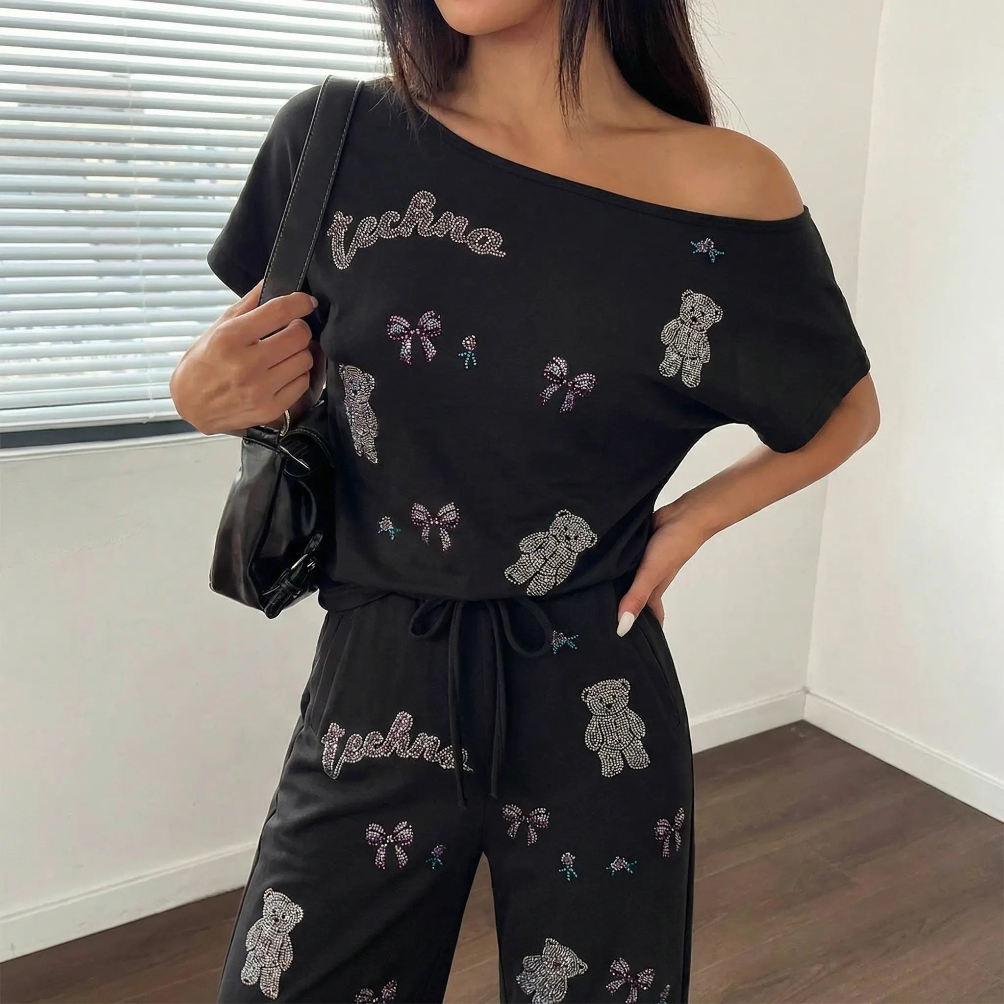 Woman wearing a black off-shoulder top and pants with embroidered designs in a room. TCH Rhinestone Teddy Bear & Bow Set