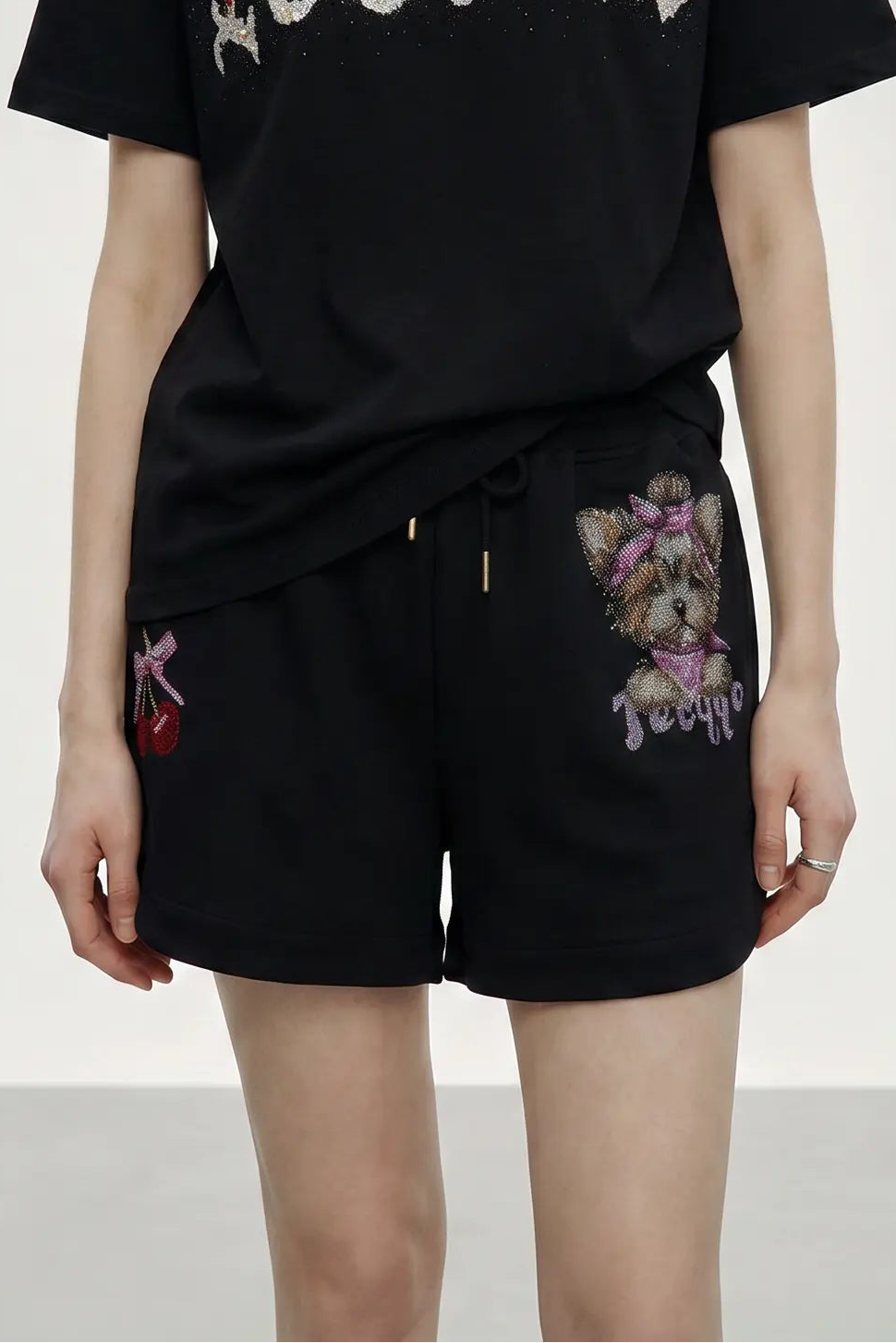 A person wearing black shorts with embroidered dog and cherry motifs, paired with a black top, shown from the chest to the upper thighs against a plain light background. TCH Rhinestone Teddy Bear Cherry Graphic Sweat Shorts