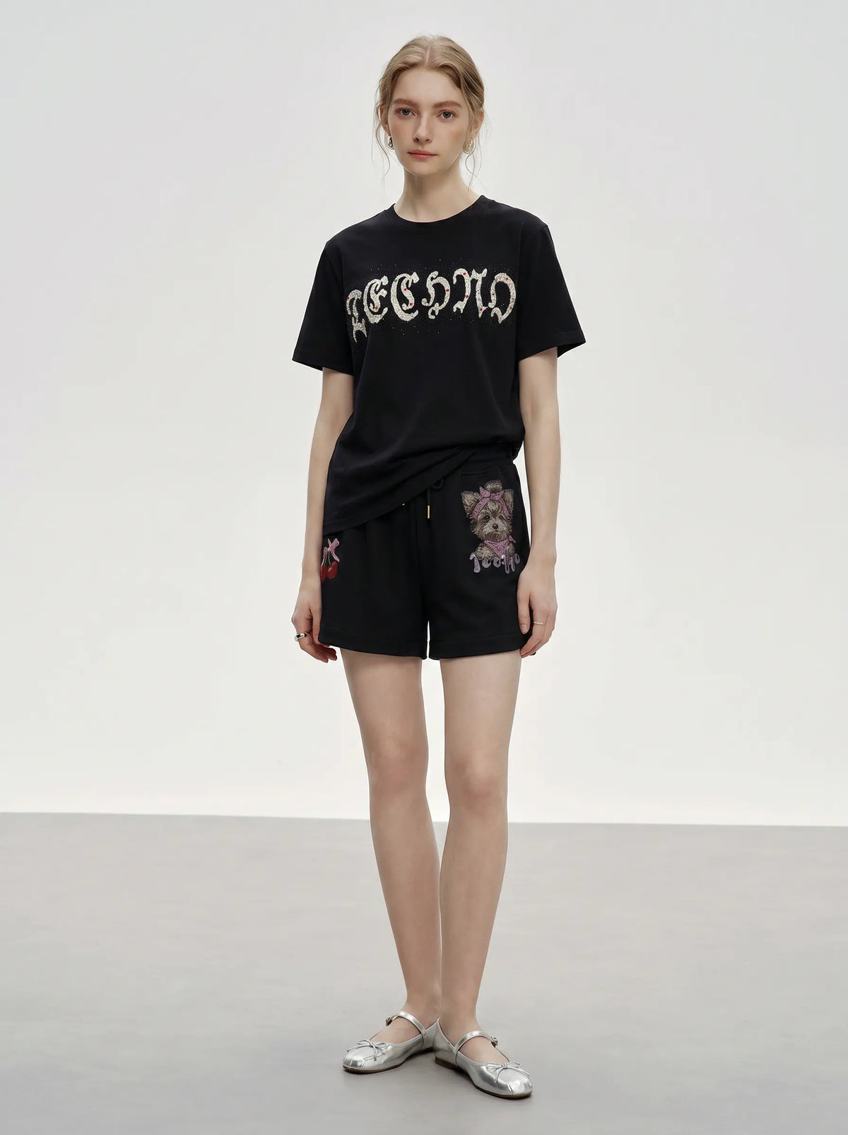 A model stands in a studio wearing a black short-sleeve T-shirt with a white gothic-style graphic print and black shorts featuring colorful floral embroidery. She is wearing silver flat shoes against a neutral light gray background. TCH Rhinestone Teddy Bear Cherry Graphic Sweat Shorts