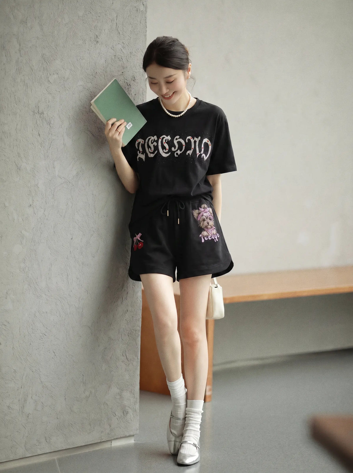A woman stands indoors against a textured gray wall, wearing a black graphic T-shirt and black shorts with floral embroidery, white socks, and white sneakers, while holding a green notebook. TCH Rhinestone Teddy Bear Cherry Graphic Sweat Shorts