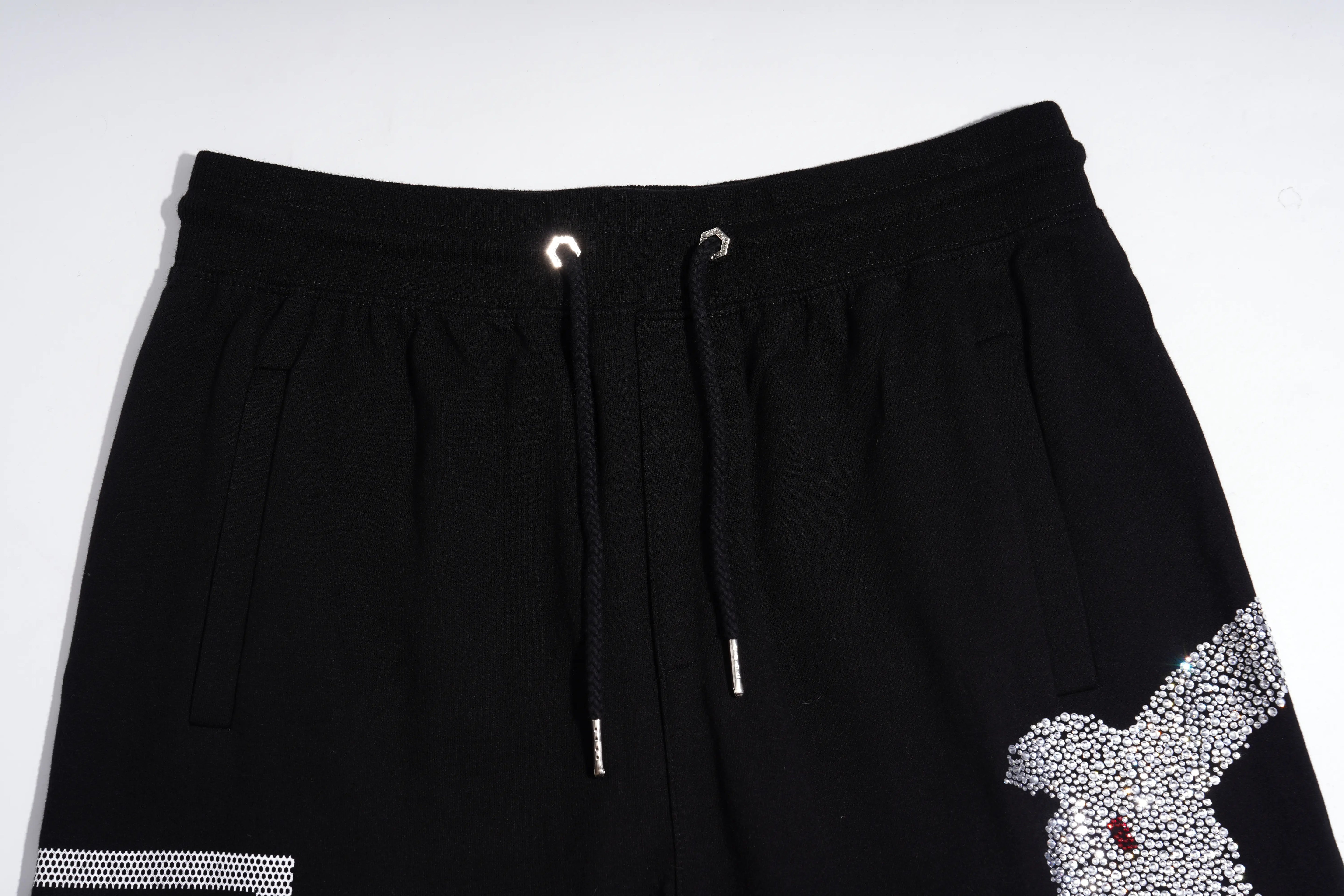 Black drawstring shorts featuring a textured white and silver bird graphic on the lower right leg, a white striped detail on the lower left leg, and silver metal eyelets on the drawstrings. TCH Rhinestone Teddy Bear Graphic Black Jogger Pants