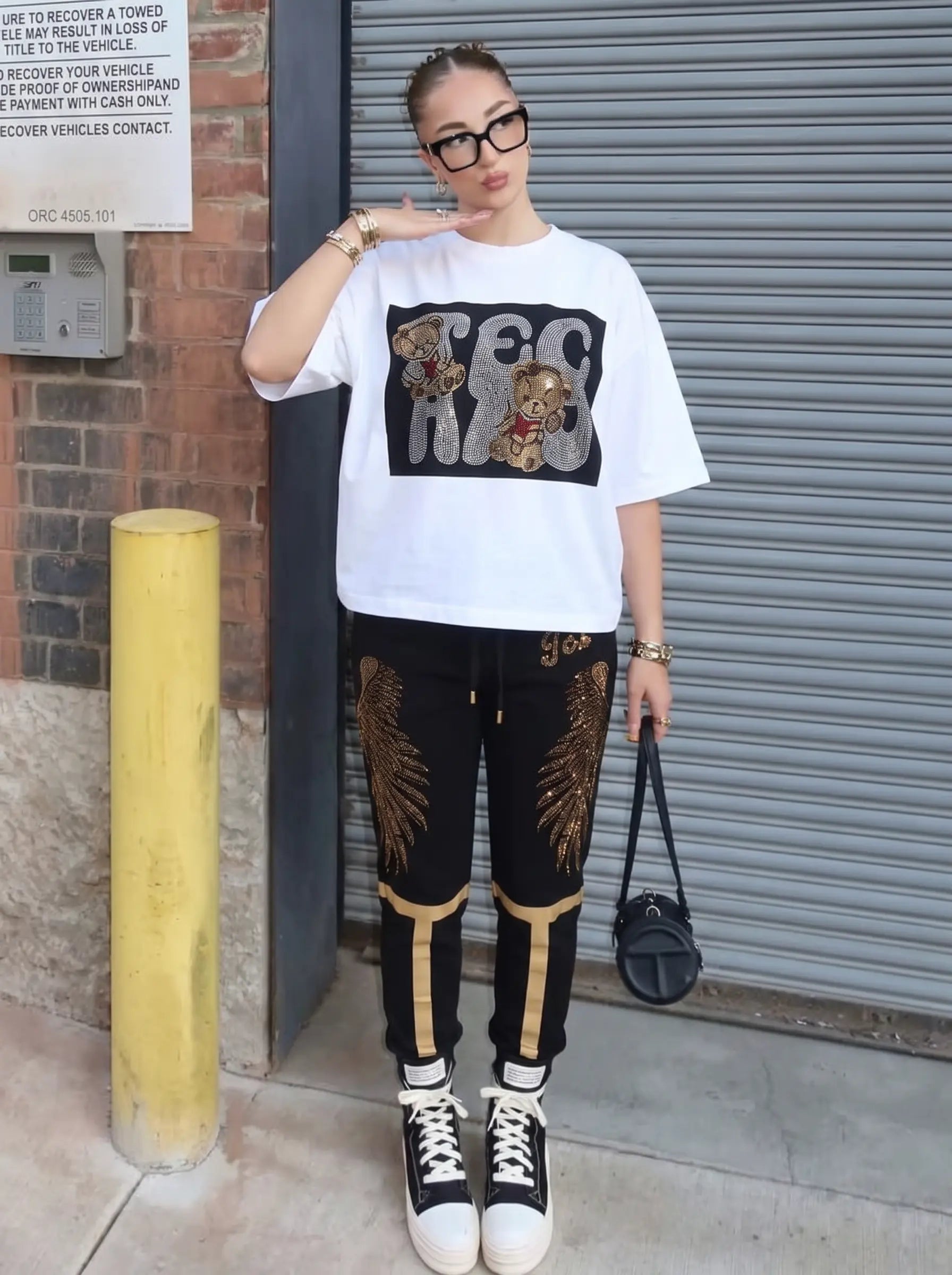 A woman stands outdoors in front of a gray metal garage door and brick wall, wearing an oversized white graphic T-shirt with a black chest print, black pants with gold wing and stripe graphics, black sneakers, and carrying a small black handbag. TCH Rhinestone Teddy Bear Graphic White T-Shirt