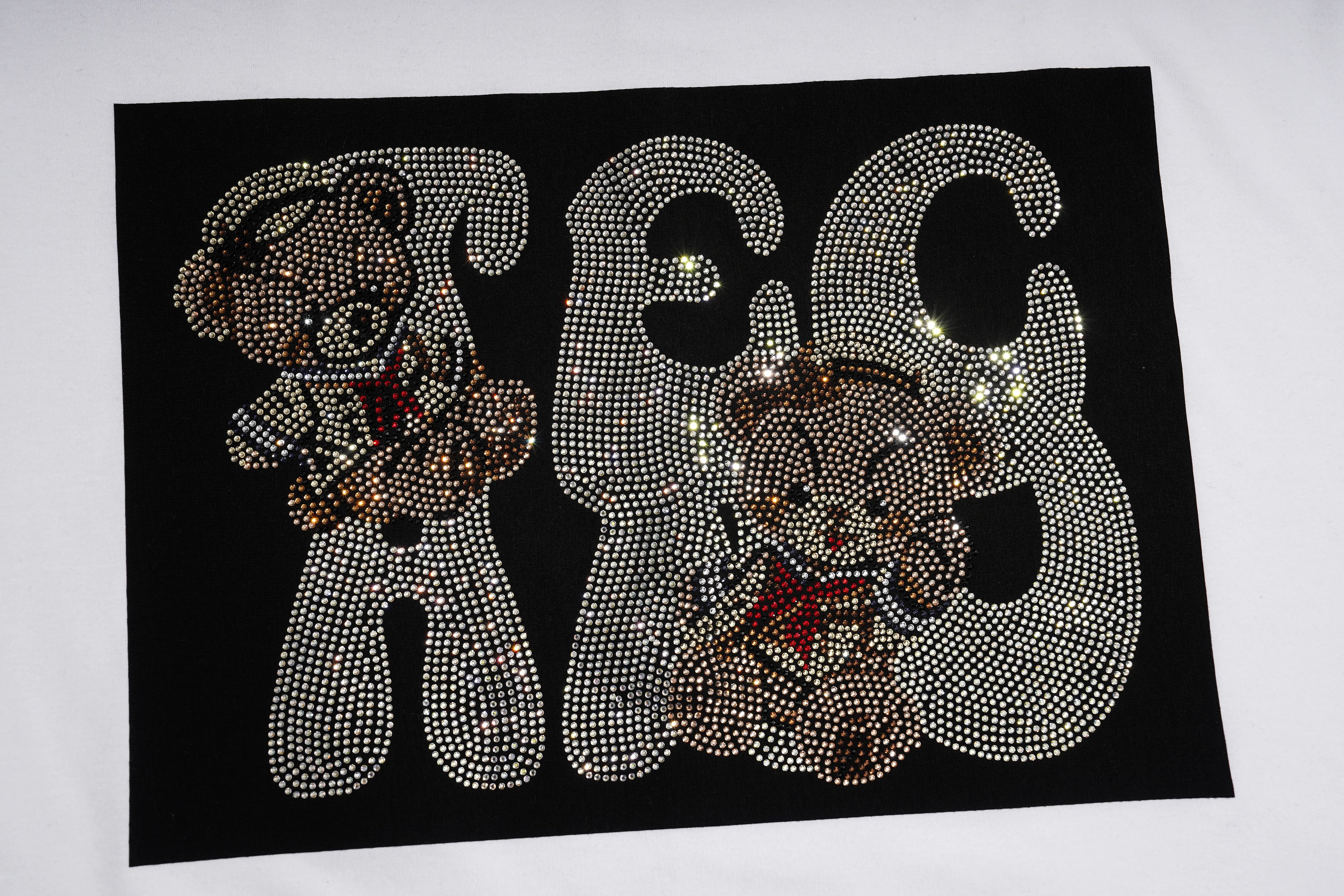 A black rectangular patch or applique featuring large outlined letters and teddy bear motifs covered in rhinestones, shown on a white background. TCH Rhinestone Teddy Bear Graphic White T-Shirt