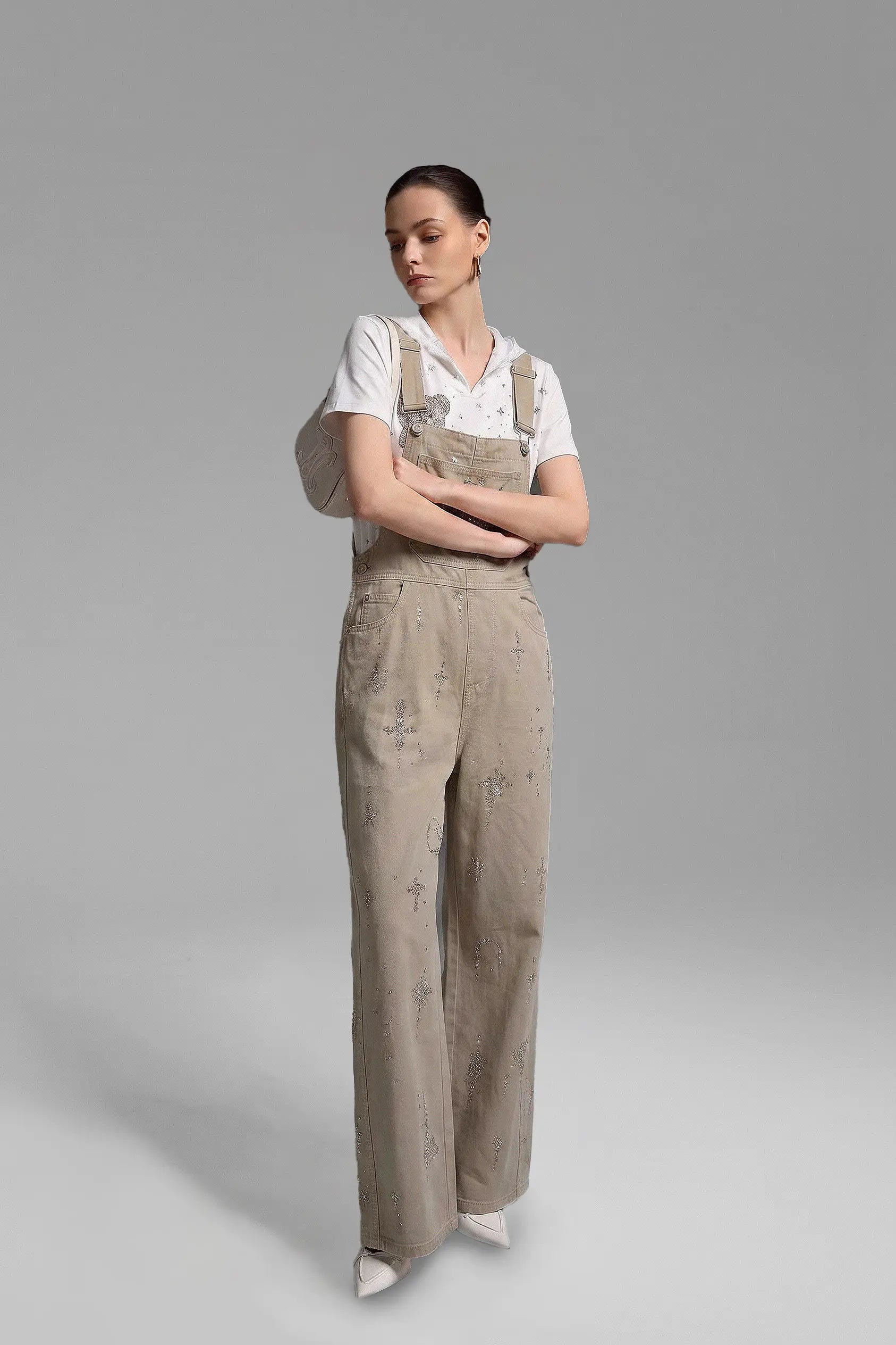 A model stands against a neutral gray studio background wearing beige wide-leg overall pants with a subtle distressed print, styled with a white short-sleeve collared shirt and white pointed shoes. TCH Rhinestone Teddy Bear Lace Hooded T-Shirt
