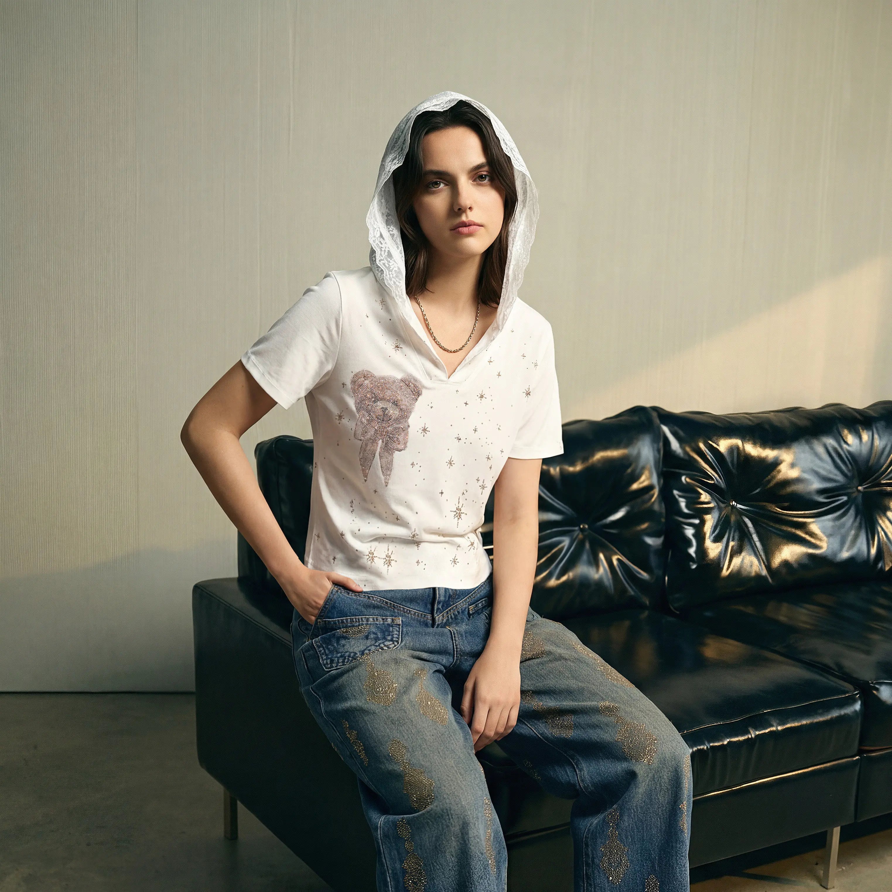 A woman wearing a white graphic t-shirt with a pinkish-brown abstract print, blue jeans, and a white lace-trimmed hood sits on a black leather couch against a textured beige wall. TCH Rhinestone Teddy Bear Lace Hooded T-Shirt