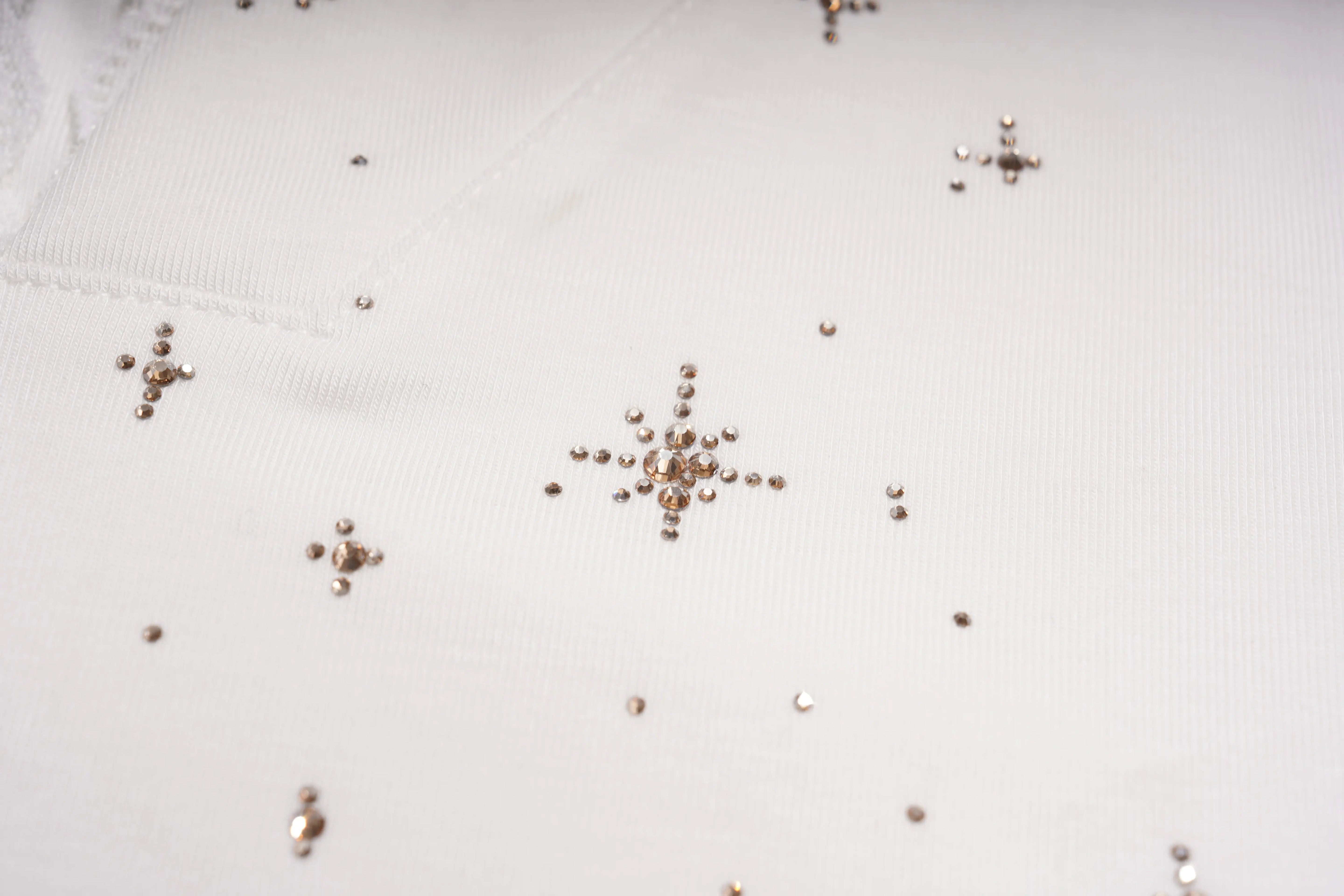 Close-up of a white fabric surface decorated with scattered metallic rose gold rhinestone snowflake and star motifs. TCH Rhinestone Teddy Bear Lace Hooded T-Shirt