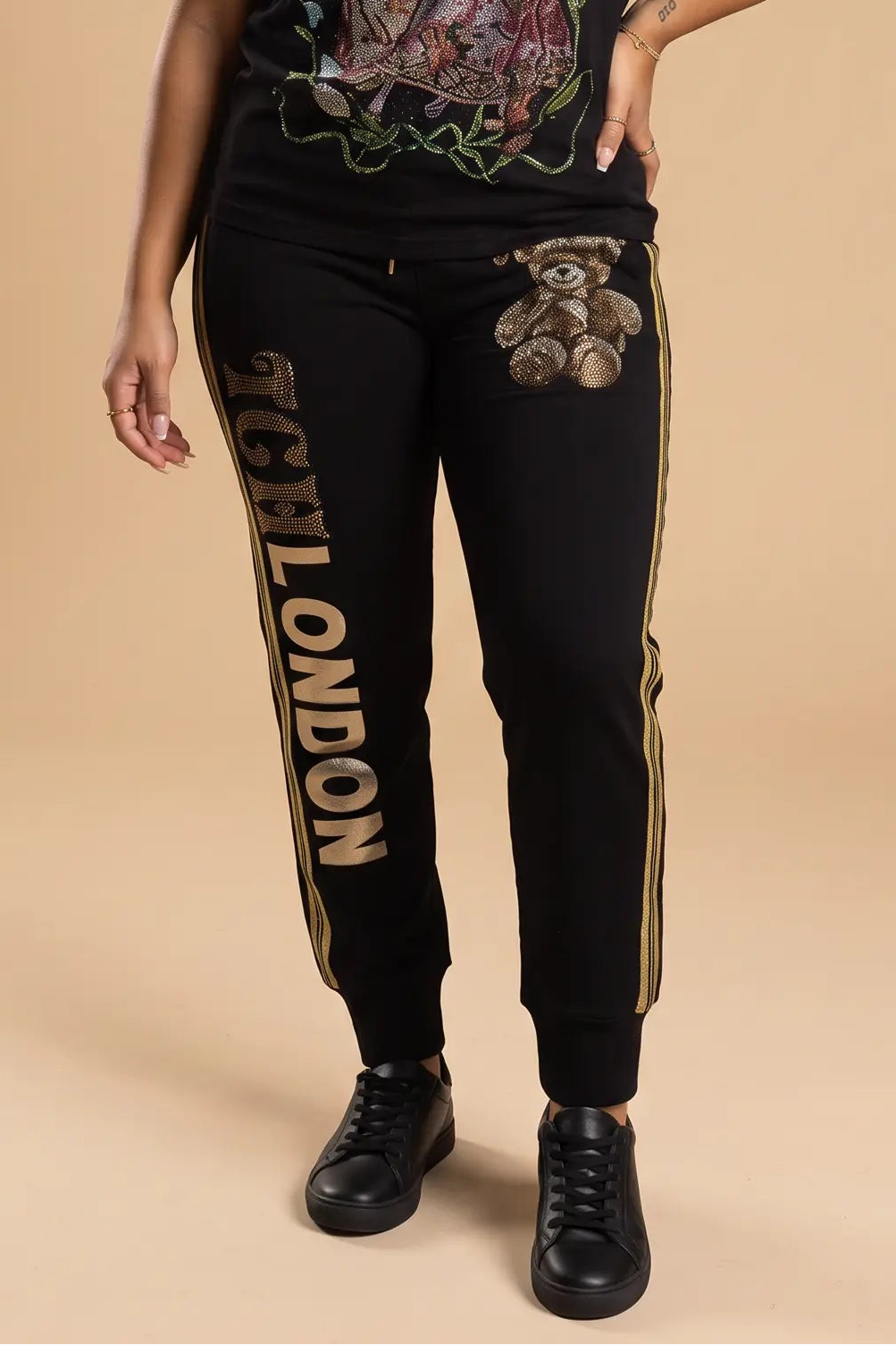 A woman modeling a black graphic t-shirt and matching black sweatpants with gold and white details, standing against a solid beige studio background. TCH Rhinestone Teddy Bear London Graphic Black Jogger Pants