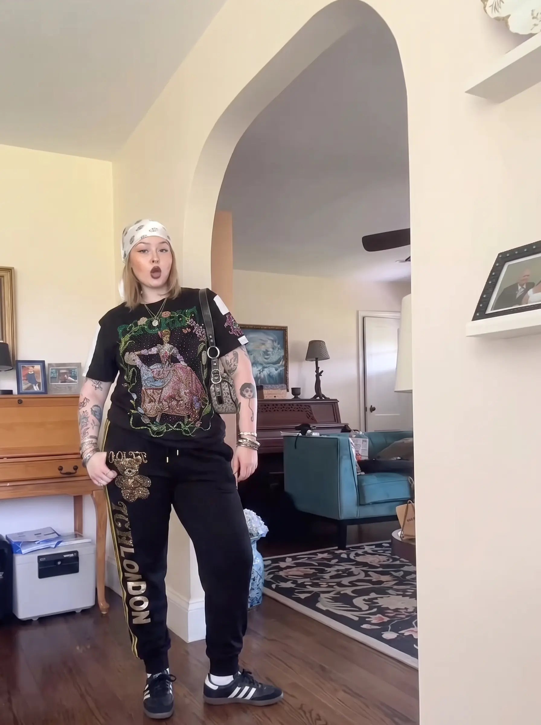 A person stands indoors wearing a black graphic T-shirt with a large multicolor front print, black sweatpants with gold lettering, a white headband, and black-and-white sneakers. The photo is taken in a home interior with an arched doorway, wood floor, furniture, and a teal chair in the background. TCH Rhinestone Teddy Bear London Graphic Black Jogger Pants