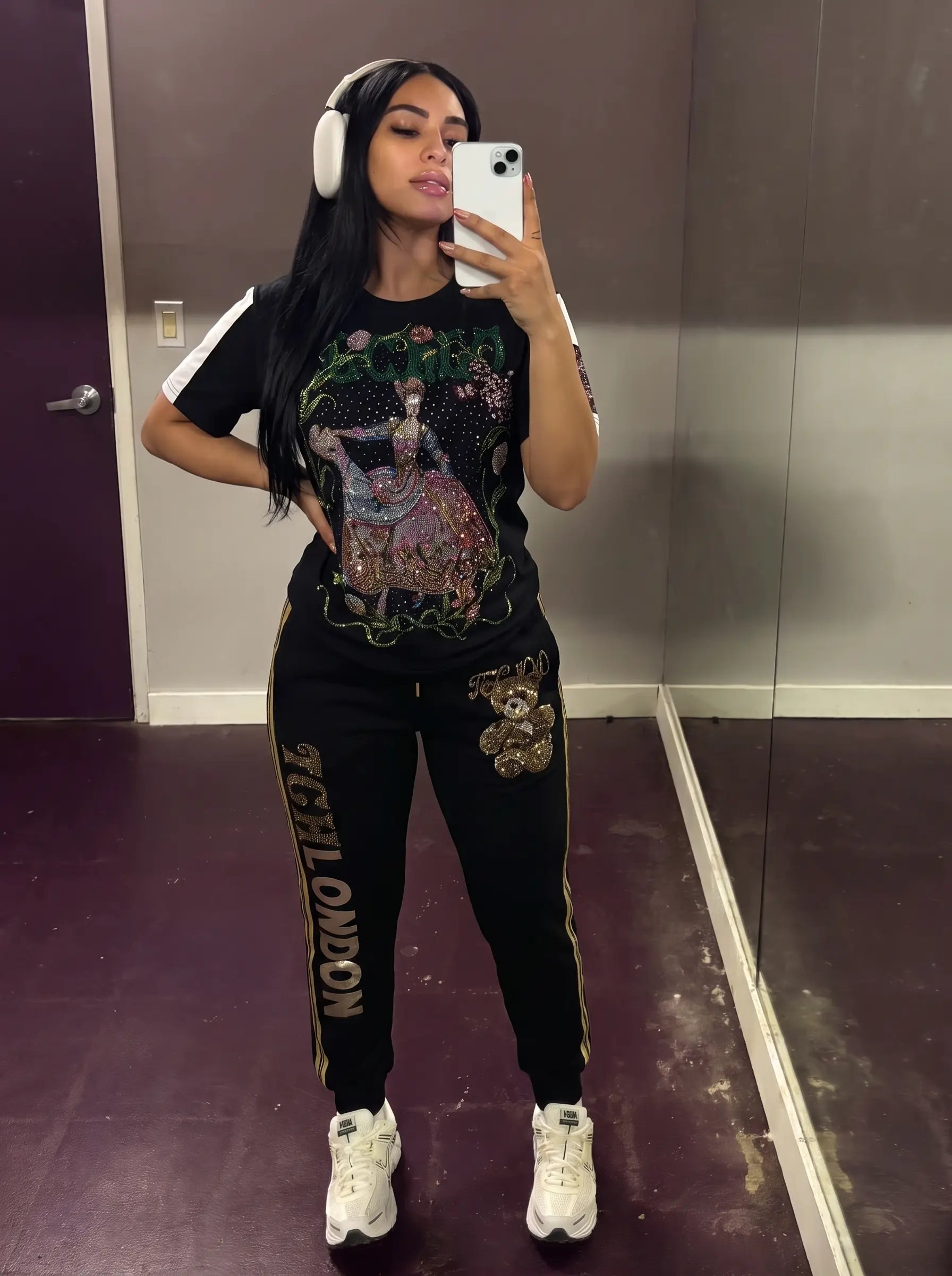 A woman takes a mirror selfie wearing a matching black graphic t-shirt and sweatpants set. The sweatpants feature gold piping and 'NYJON' text down the leg, with a teddy bear patch on the thigh. TCH Rhinestone Teddy Bear London Graphic Black Jogger Pants