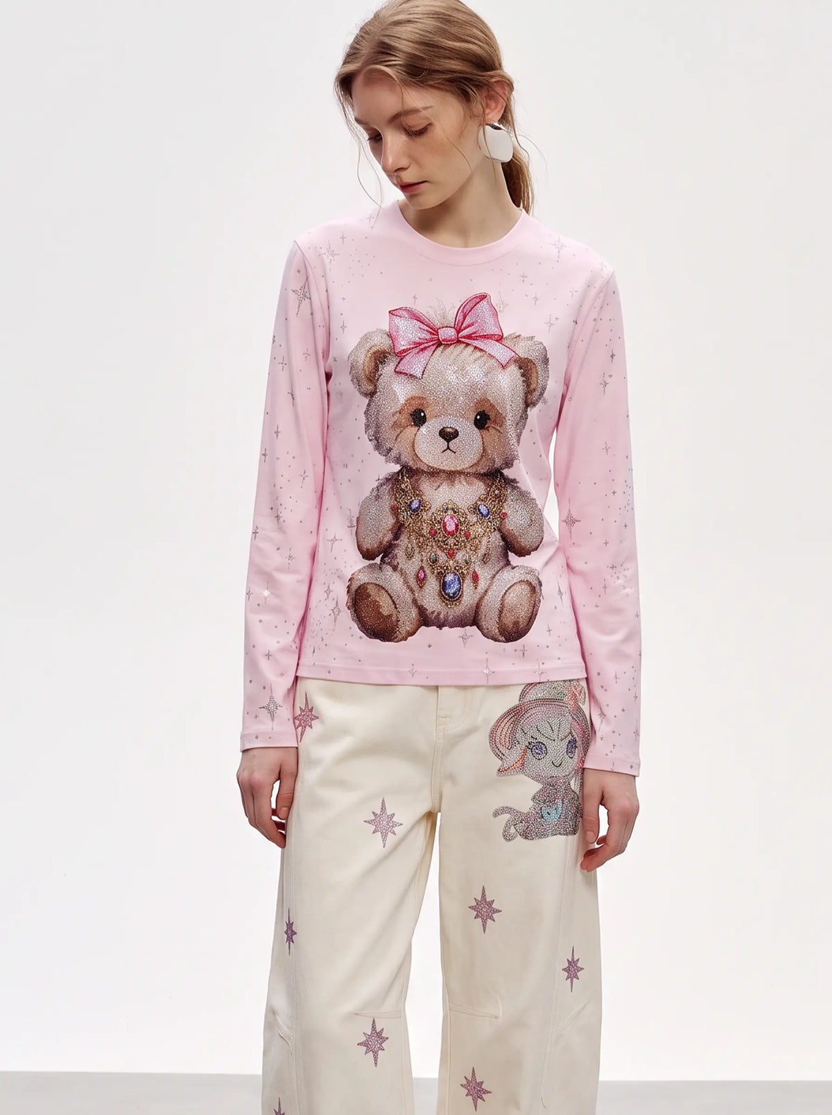 A model wearing a light pink long-sleeve sweatshirt with a large teddy bear graphic on the front, paired with cream-colored pants featuring star prints. TCH Rhinestone Teddy Bear Pink Long Sleeve T-Shirt