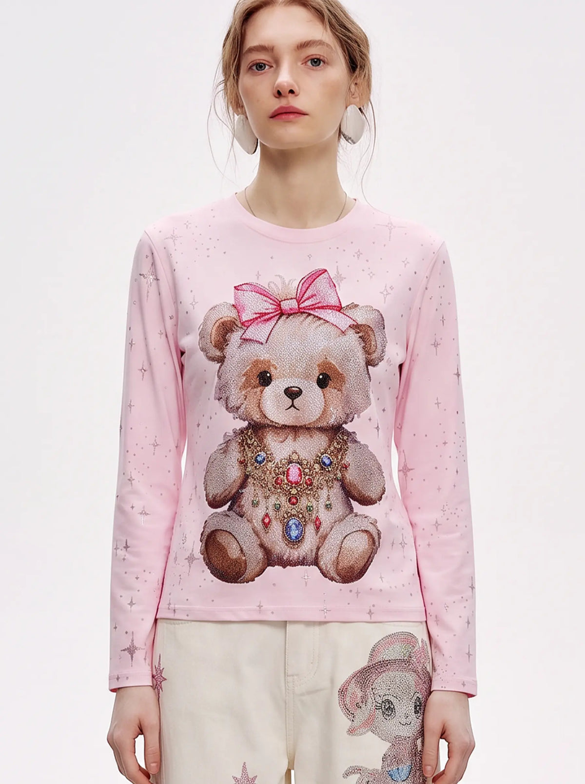 A model wearing a light pink long-sleeve crewneck sweatshirt featuring a large teddy bear graphic on the front with a pink bow and jeweled collar, paired with cream-colored pants. TCH Rhinestone Teddy Bear Pink Long Sleeve T-Shirt