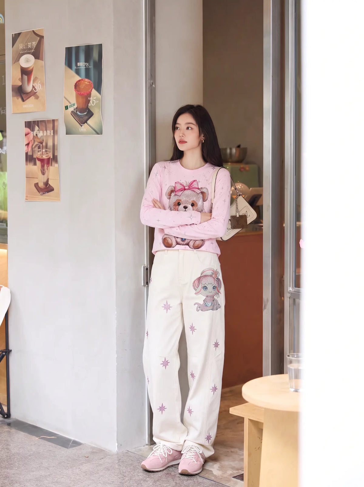 A woman stands in a doorway wearing a pink long-sleeve top and cream-colored sweatpants with cartoon bear graphics and star prints, paired with pink sneakers. TCH Rhinestone Teddy Bear Pink Long Sleeve T-Shirt
