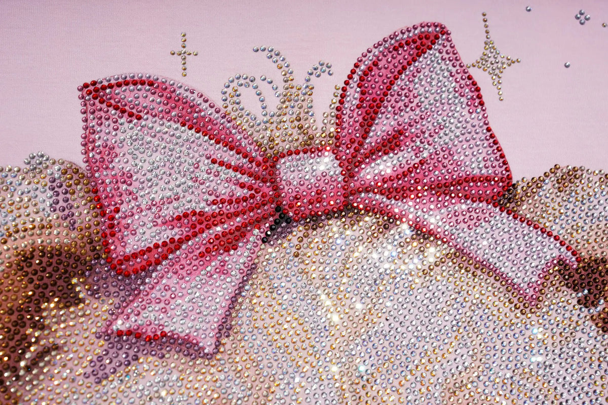 A close-up of a pink rhinestone bow embellishment with a matching pink and silver beaded crown, set against a light pink background with small gold glitter star accents. TCH Rhinestone Teddy Bear Pink Long Sleeve T-Shirt