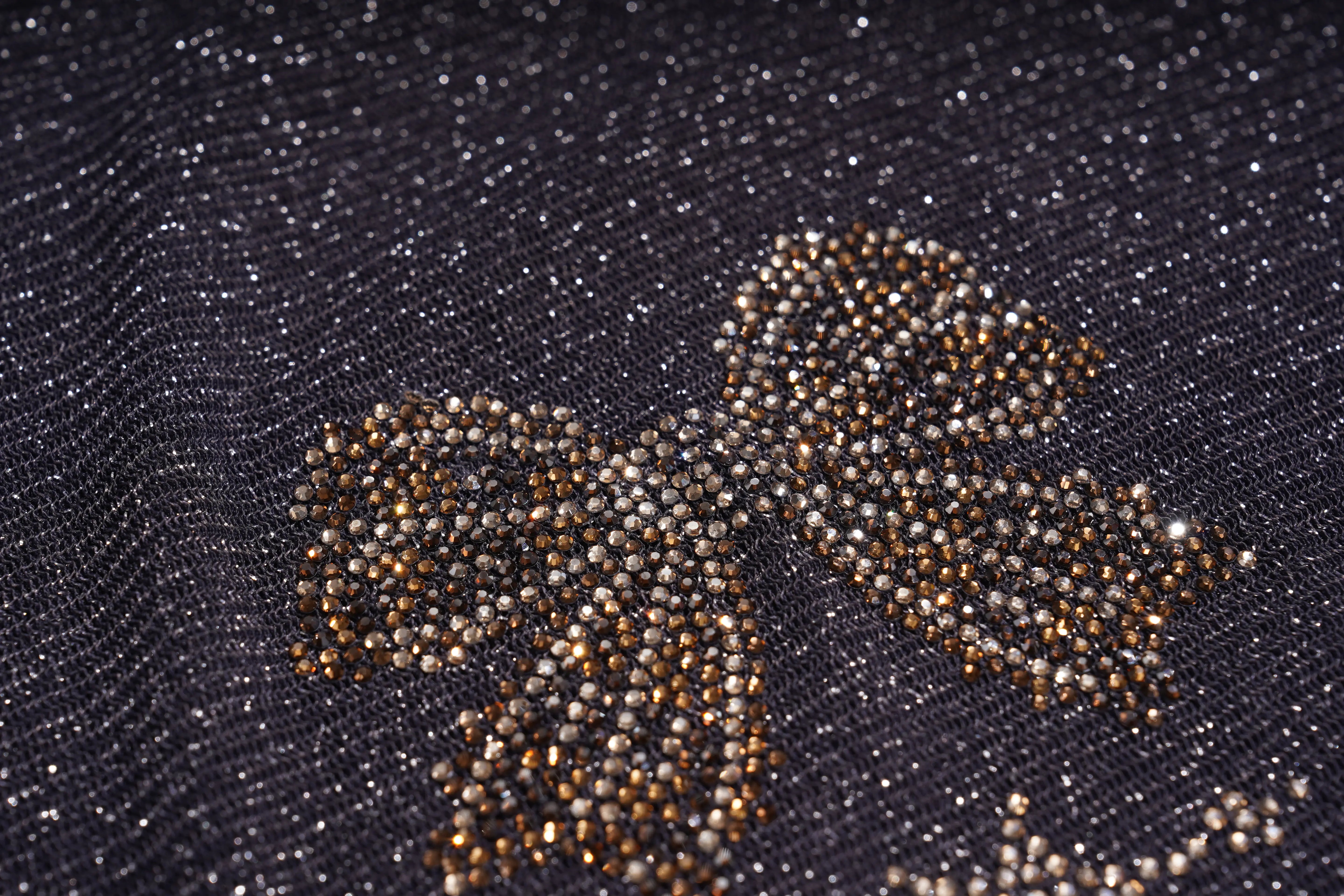 Close-up of a dark textured fabric with a decorative bow motif made from small metallic beads in silver, gold, and bronze tones. TCH Rhinestone Teddy Bear Sheer Mesh Long Sleeve Top