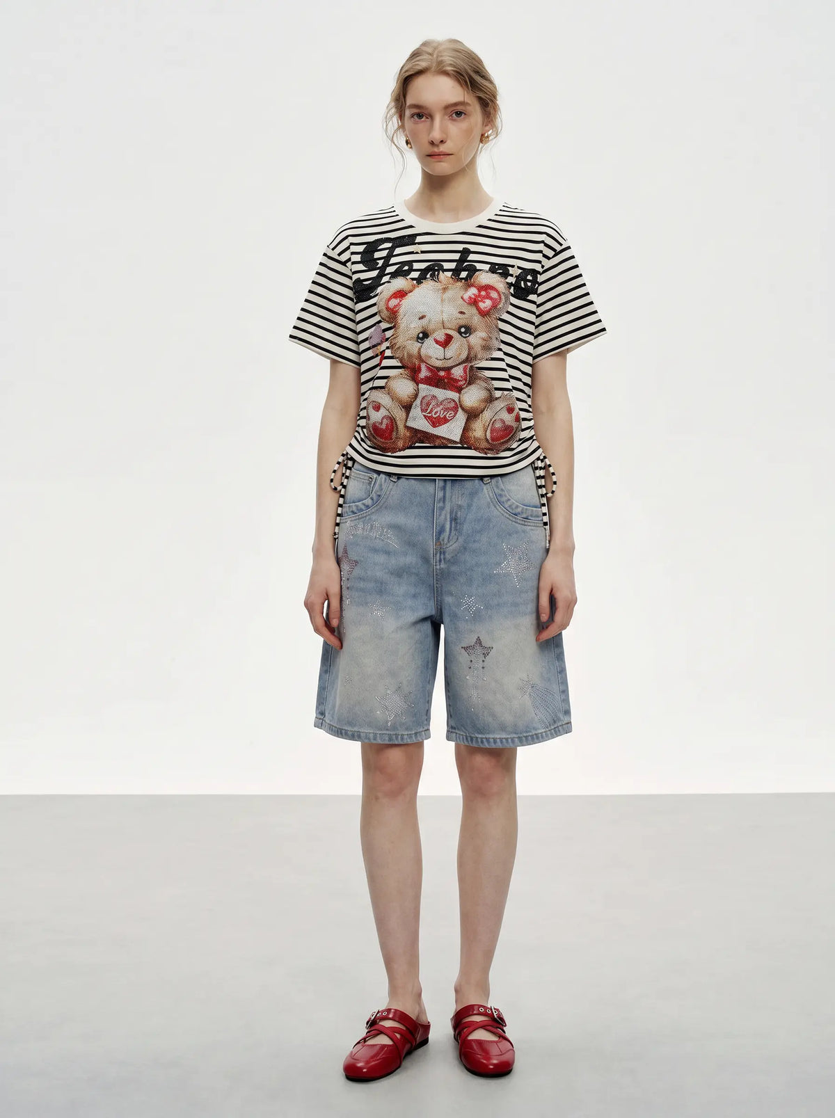 A model stands against a light gray studio background wearing a black and white striped graphic t-shirt with a teddy bear print, light-wash denim shorts, and red loafers. TCH Rhinestone Teddy Bear Striped Tie-Side T-Shirt