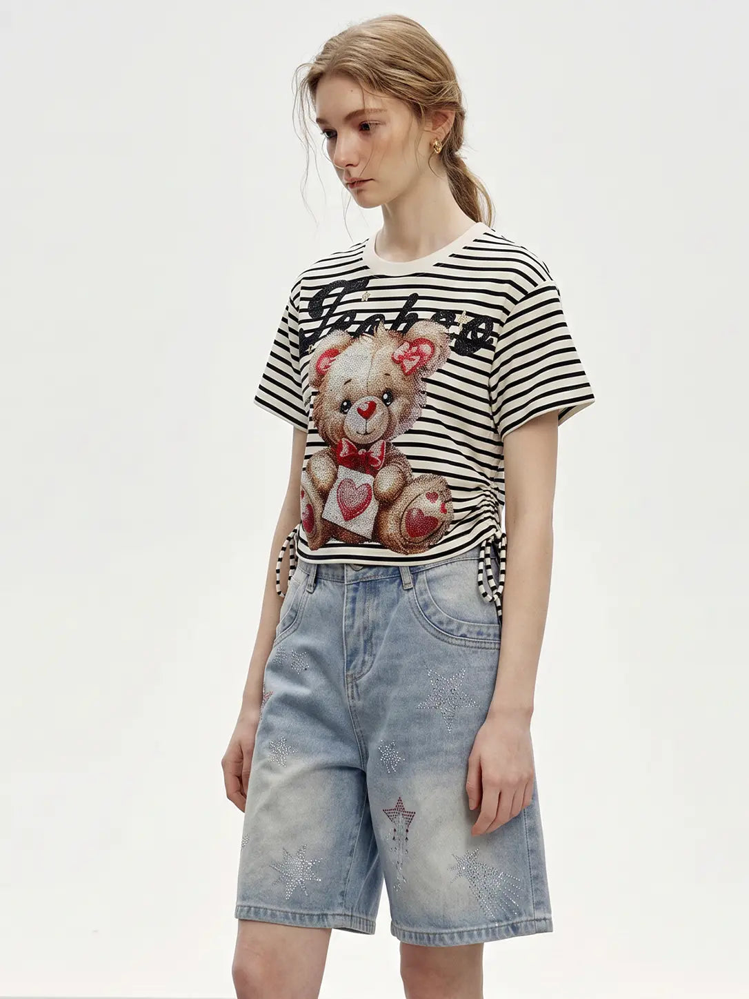A woman modeling a black and white striped t-shirt with a teddy bear graphic holding a heart, paired with light blue denim shorts, against a plain white studio background. TCH Rhinestone Teddy Bear Striped Tie-Side T-Shirt