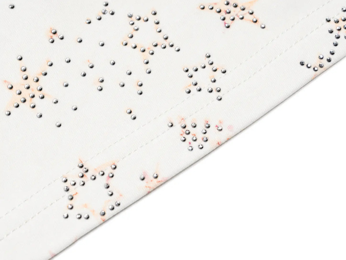 Close-up of a white fabric surface with scattered metallic silver studs and a few faint peach-pink embroidered or printed accents, photographed on a white background. TCH Rhinestone Teddy Bear Techno Raglan T-Shirt