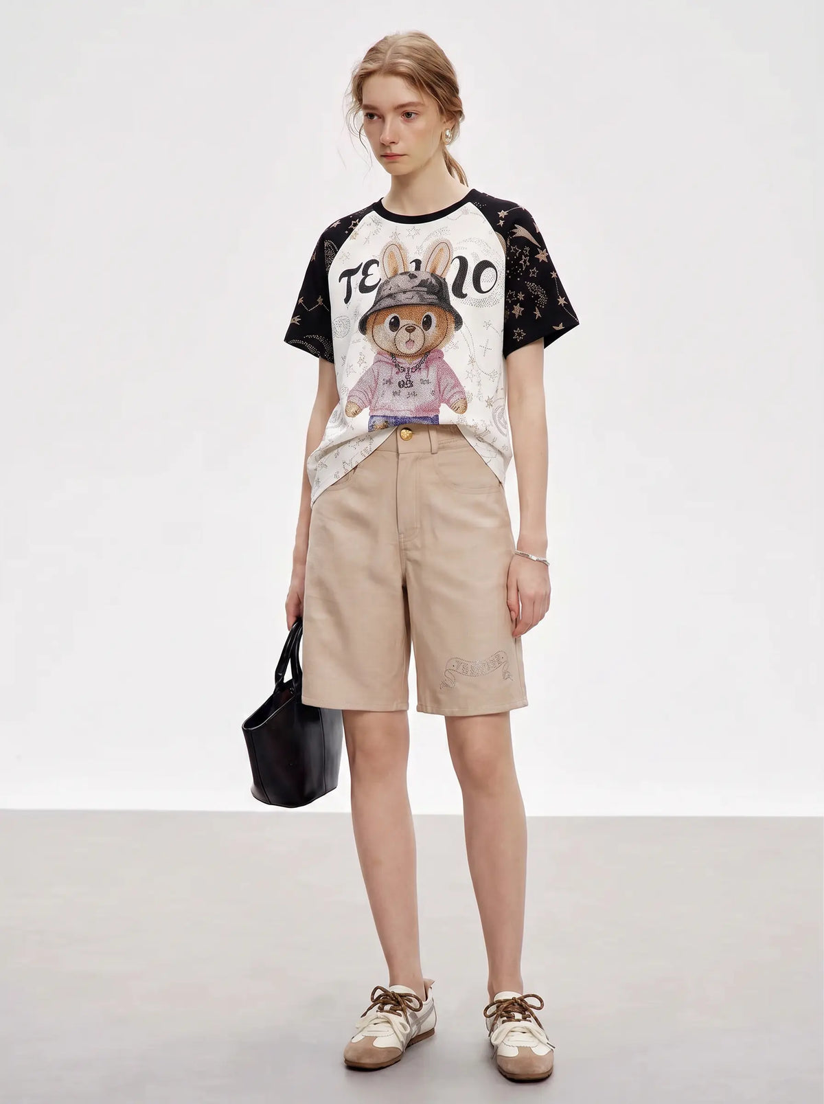 A model stands in a studio wearing a graphic T-shirt with a rabbit illustration and black floral sleeves, paired with beige shorts, white sneakers, and holding a black handbag. TCH Rhinestone Teddy Bear Techno Raglan T-Shirt