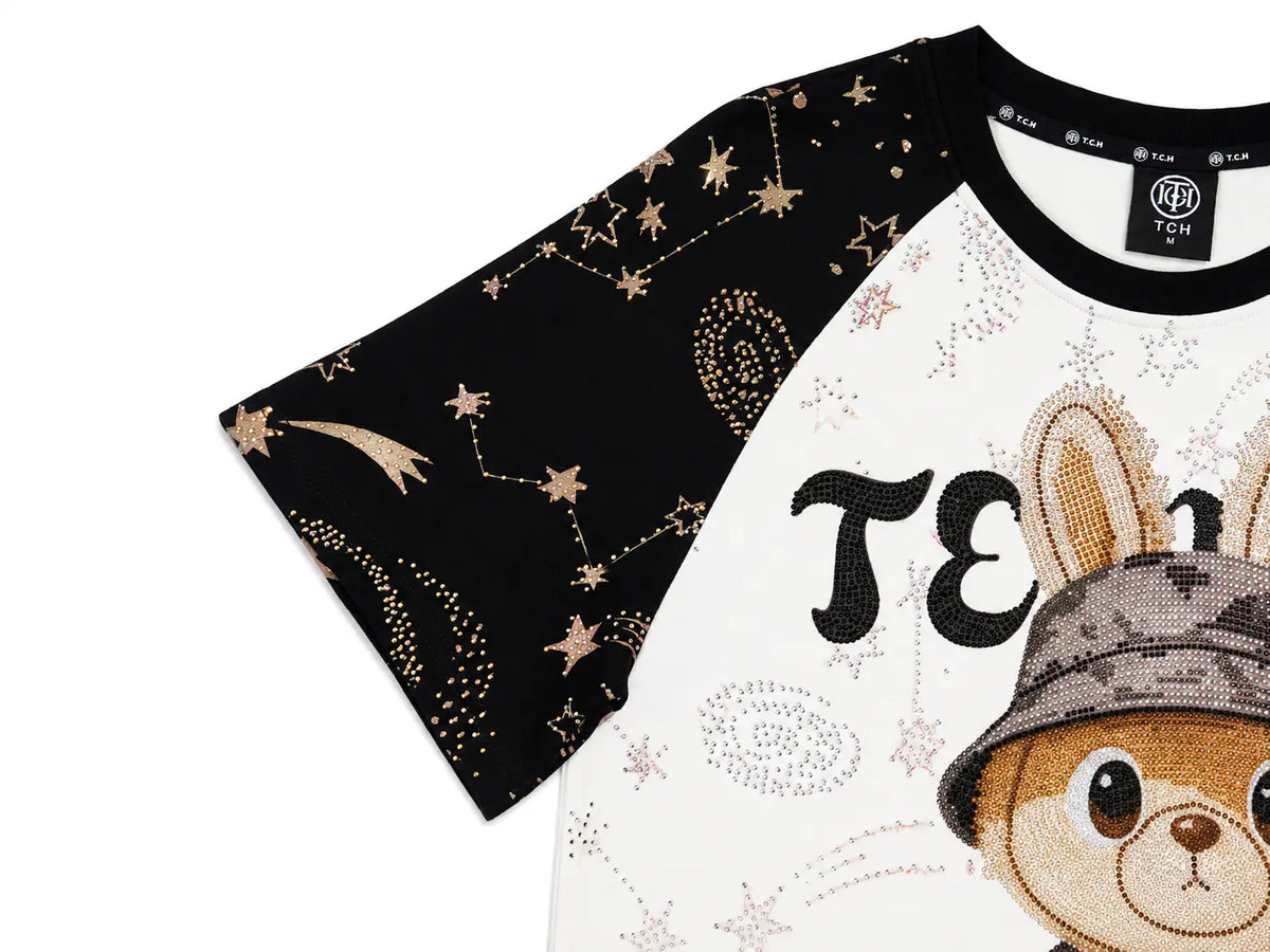 A close-up of a raglan-style T-shirt with a black sleeve decorated with gold stars and constellations, and a white body featuring a cartoon rabbit wearing a camouflage hat and sequined lettering. TCH Rhinestone Teddy Bear Techno Raglan T-Shirt