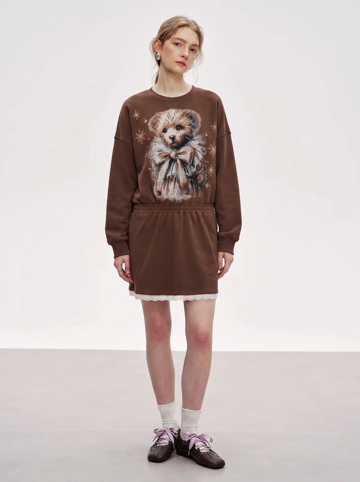 A model stands in a studio wearing a brown sweatshirt dress featuring a large teddy bear graphic on the front, styled with white socks and dark brown shoes. TCH Rhinestone Teddy Bear Techno Sweatshirt Dress