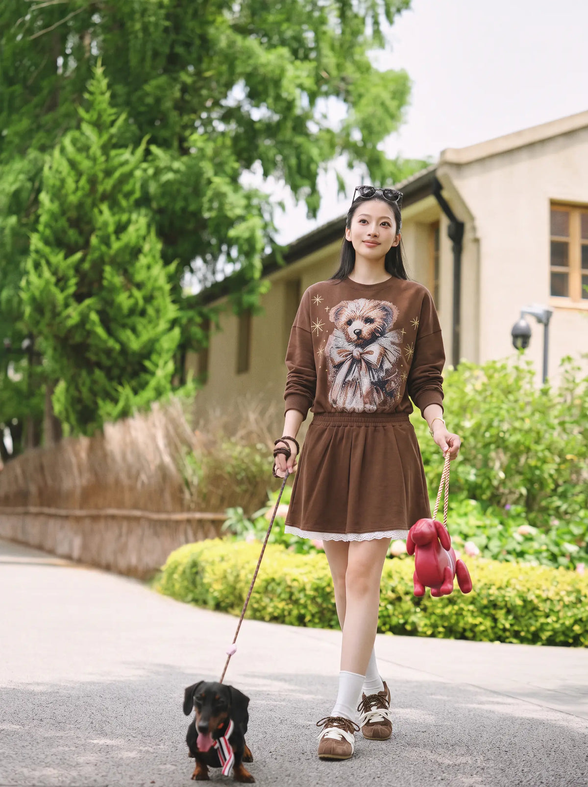 A woman walking a small black dog on a paved path outdoors, wearing a brown long-sleeve dress with a large bear graphic on the front and holding a red plush toy. TCH Rhinestone Teddy Bear Techno Sweatshirt Dress