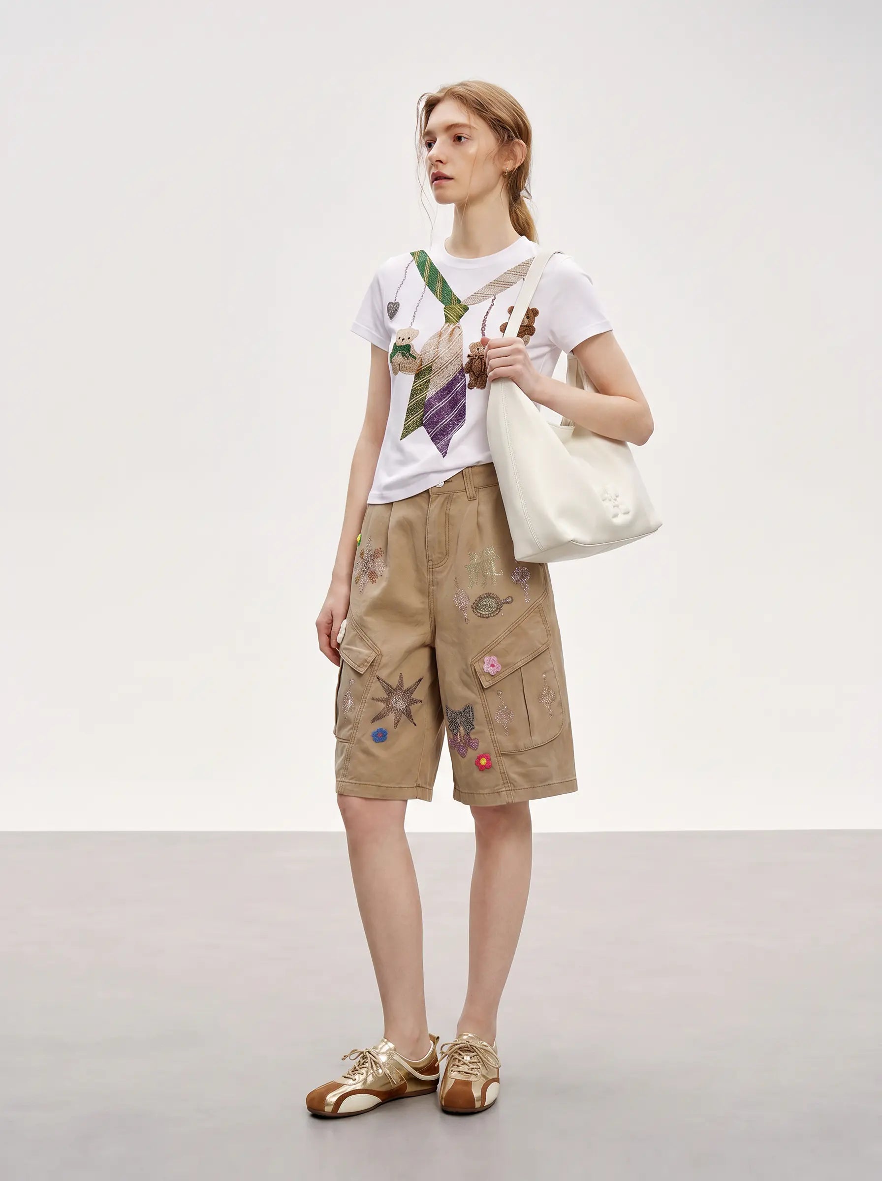 A model stands in a studio wearing a white graphic T-shirt, tan cargo shorts with colorful embroidered details, and brown loafers, while carrying a large off-white tote bag. TCH Rhinestone Teddy Bear Tie Graphic White T-Shirt