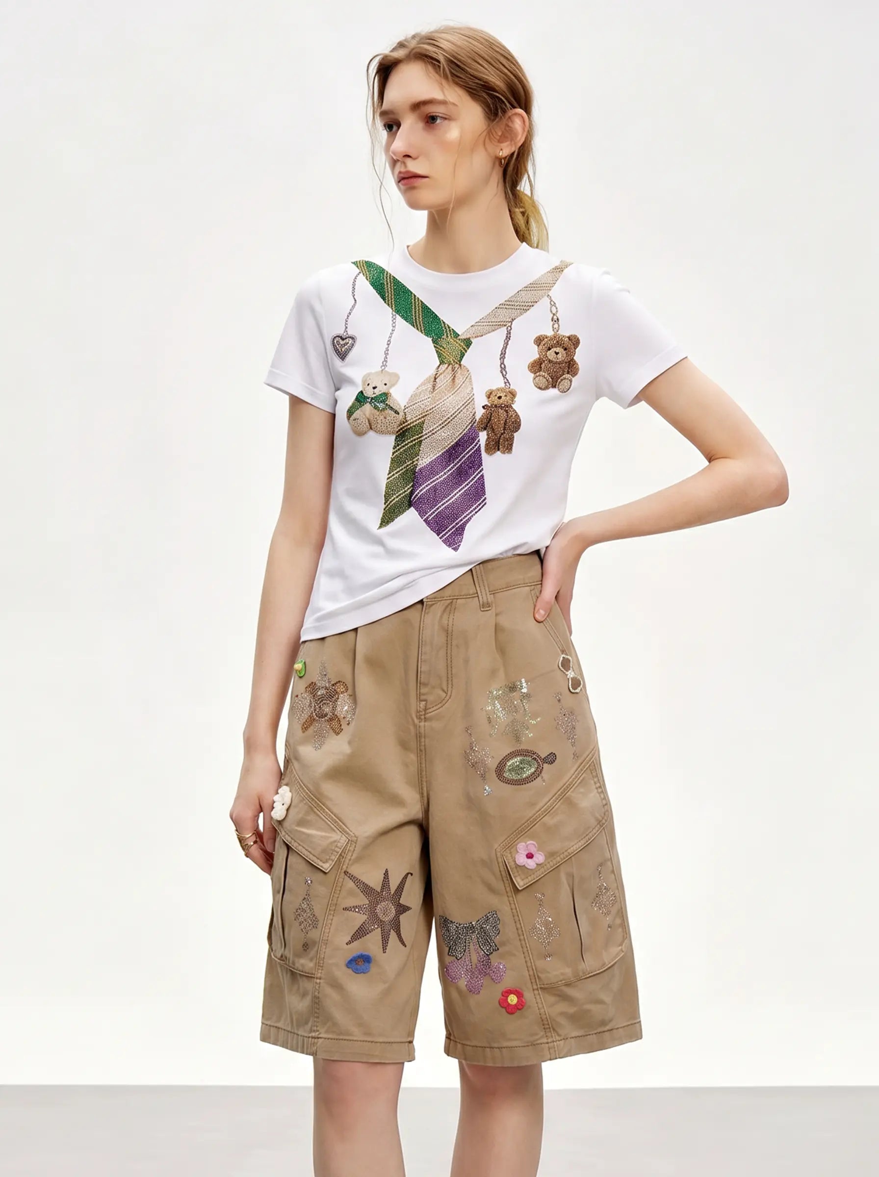 A model wearing a white graphic t-shirt with a tie and teddy bear print, paired with beige cargo shorts decorated with colorful patches, against a plain light gray background. TCH Rhinestone Teddy Bear Tie Graphic White T-Shirt