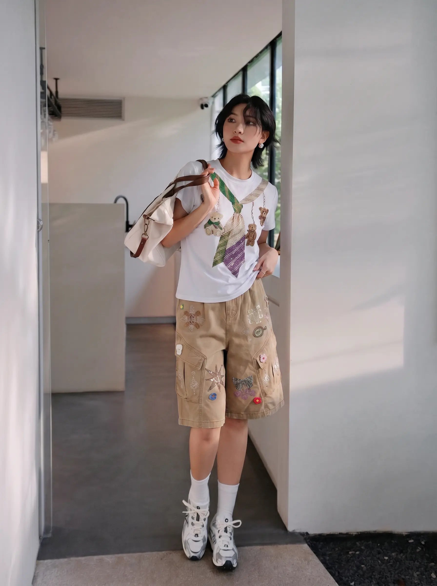 A woman stands in a modern indoor hallway wearing a white graphic T-shirt, beige cargo shorts with colorful patches, white socks, and white sneakers, while carrying a beige shoulder bag. TCH Rhinestone Teddy Bear Tie Graphic White T-Shirt