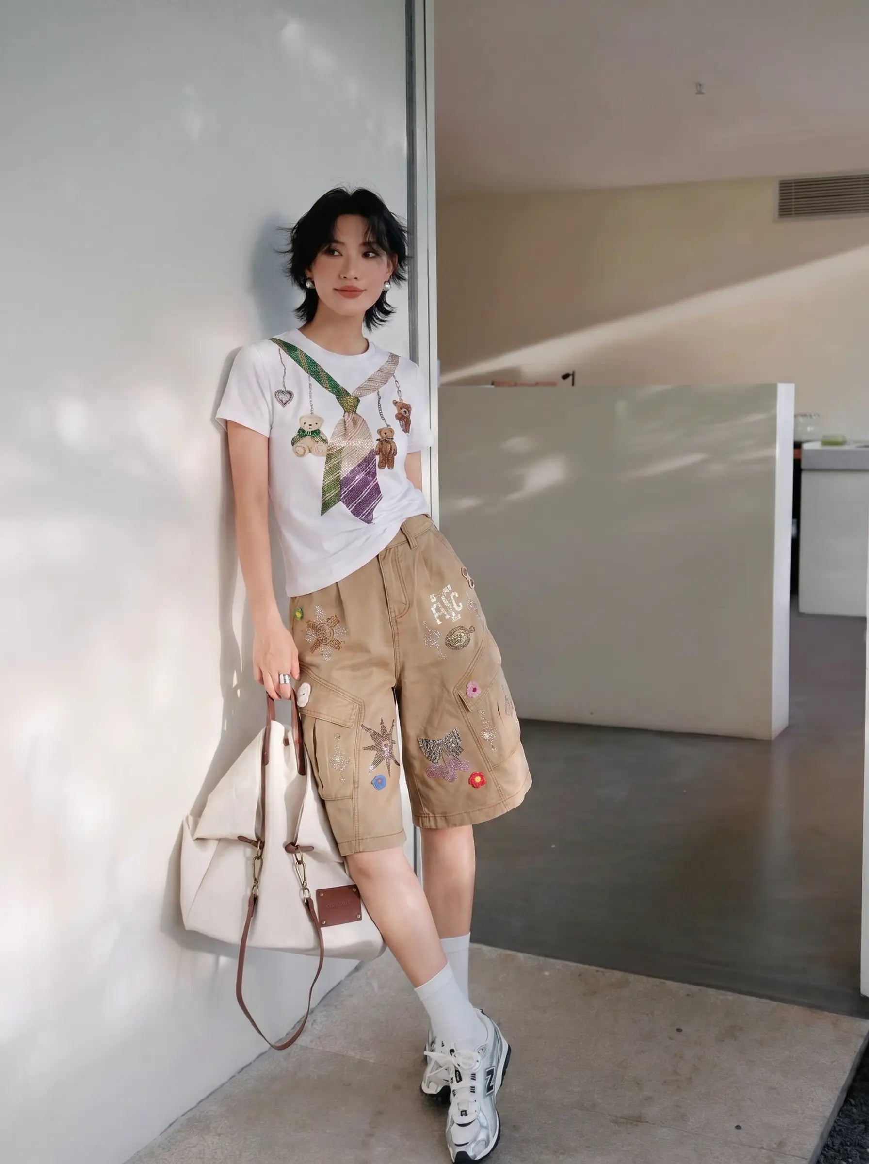 A woman stands in a modern indoor setting, leaning against a white wall. She is wearing a white graphic t-shirt, beige cargo shorts with colorful patches, white socks, and white sneakers, and holding a large cream-colored shoulder bag. TCH Rhinestone Teddy Bear Tie Graphic White T-Shirt