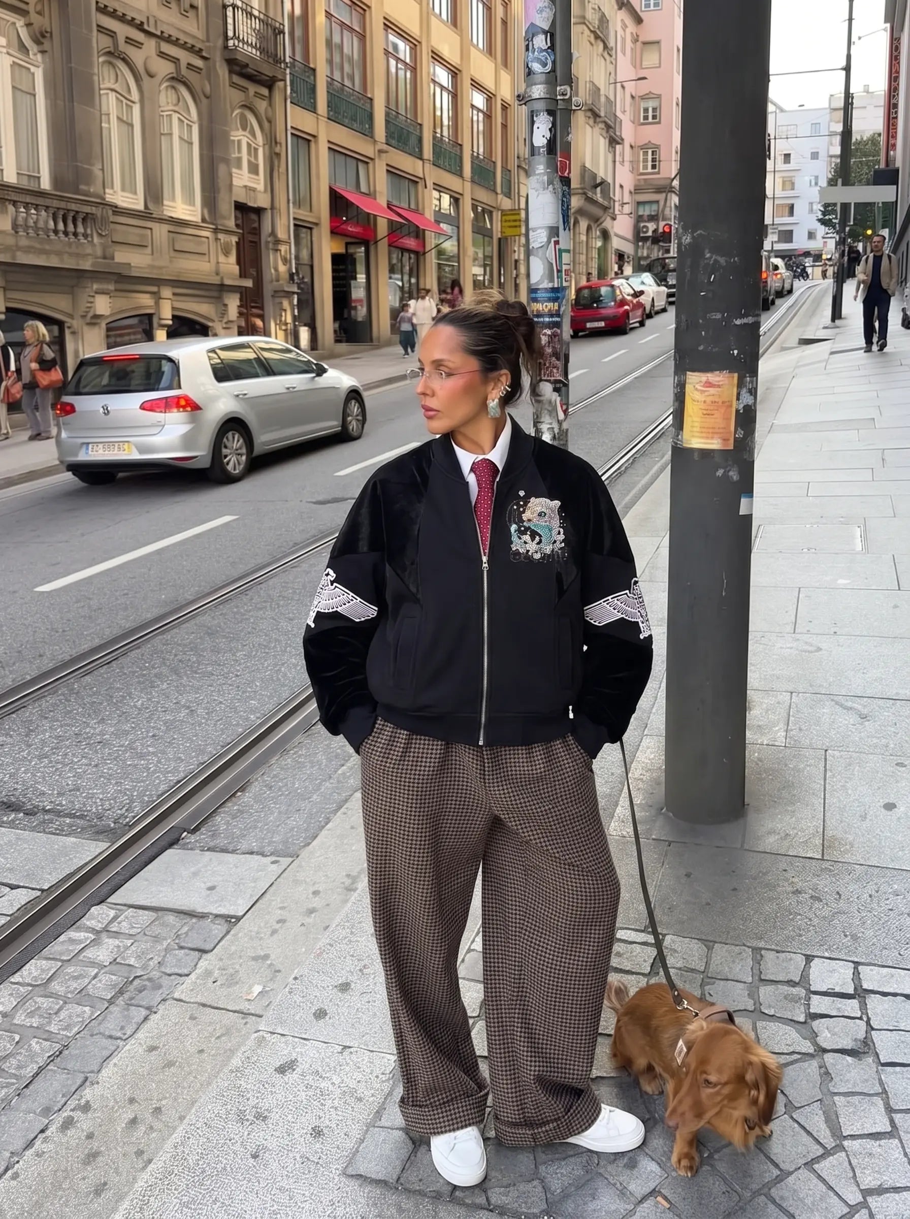A woman stands on a city street sidewalk wearing a black bomber jacket with white sleeve stripes, brown wide-leg patterned trousers, a white collared shirt, and a red tie. She is holding a leash with a small brown dog next to her, with buildings, cars, and a streetlamp in the background. TCH Rhinestone Teddy Bear Wing Sleeve Bomber Jacket
