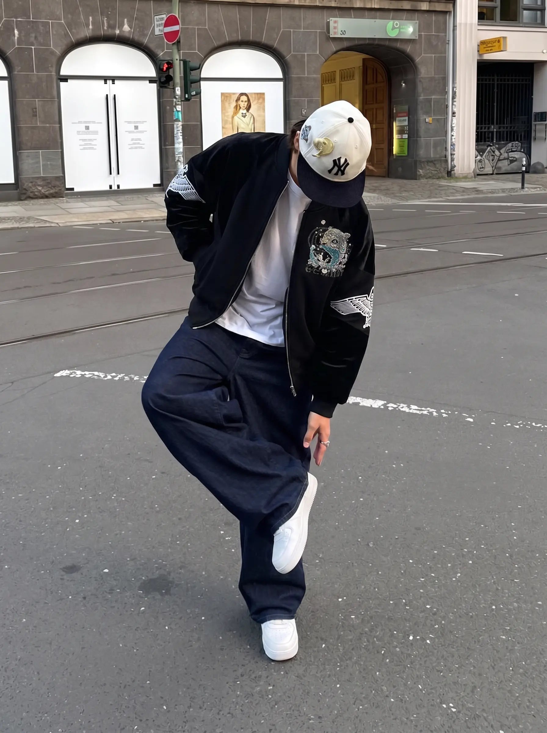 A person posing on a city street wearing a black bomber jacket with white sleeve patches, navy blue baggy sweatpants, a white t-shirt, white sneakers, and a white baseball cap with a black NY logo. TCH Rhinestone Teddy Bear Wing Sleeve Bomber Jacket