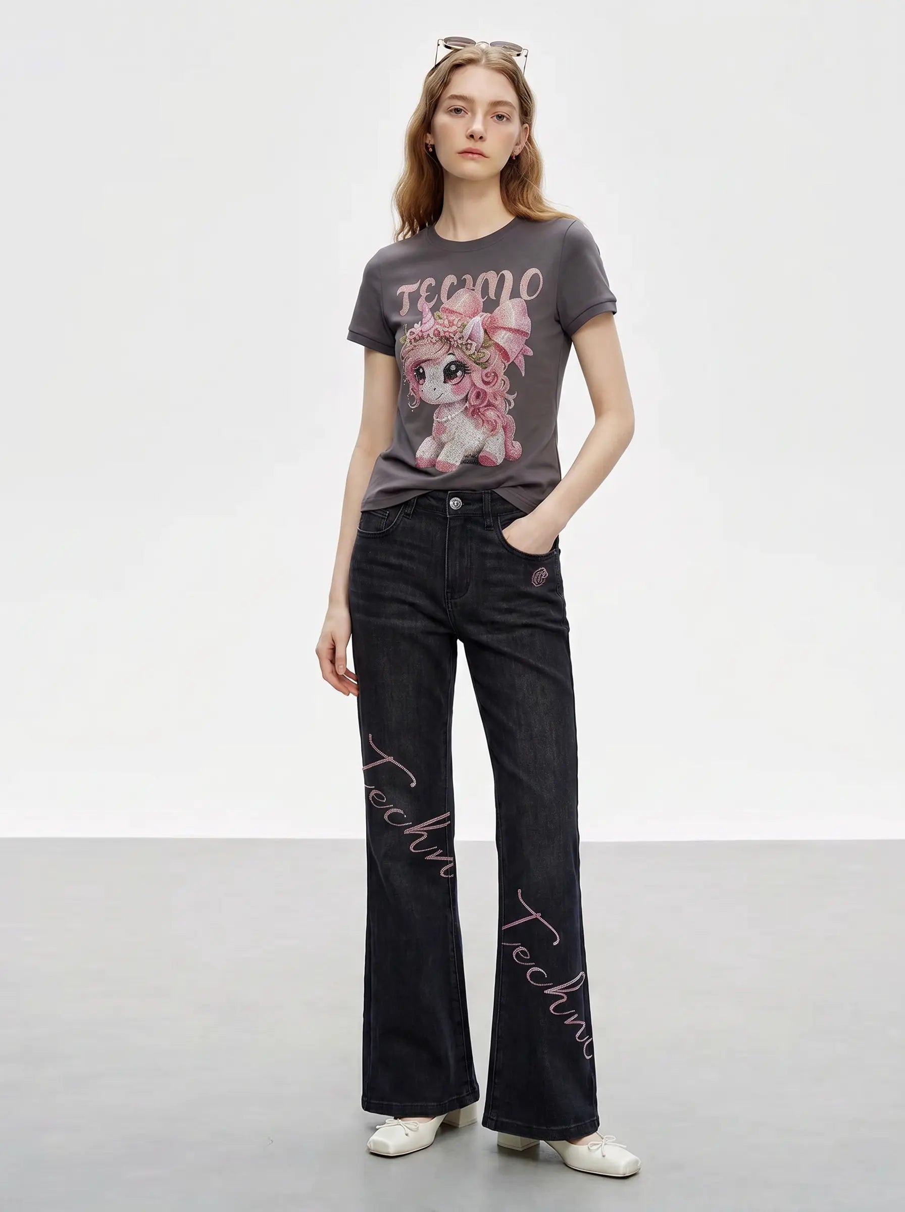 A model stands in a studio wearing a dark gray graphic t-shirt with a pink cartoon bear and the word 'teemo', paired with black wide-leg jeans featuring pink script embroidery down the front, and white sneakers. TCH Rhinestone Unicorn Graphic Charcoal T-Shirt