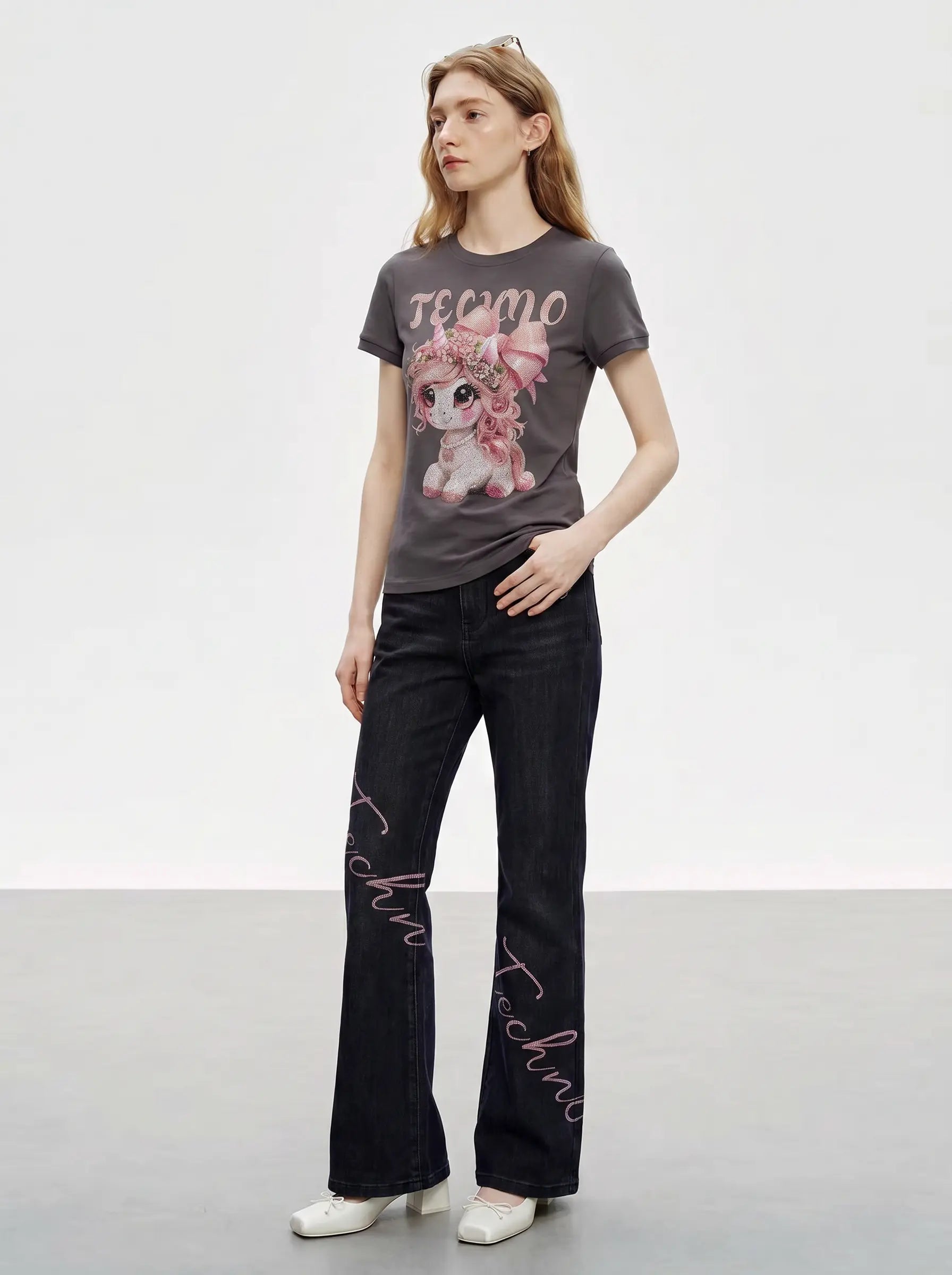 A woman modeling a dark gray graphic t-shirt with a cartoon character and the word 'REMO', paired with dark wide-leg jeans featuring pink script text down the legs, and white shoes, set against a plain studio background. TCH Rhinestone Unicorn Graphic Charcoal T-Shirt