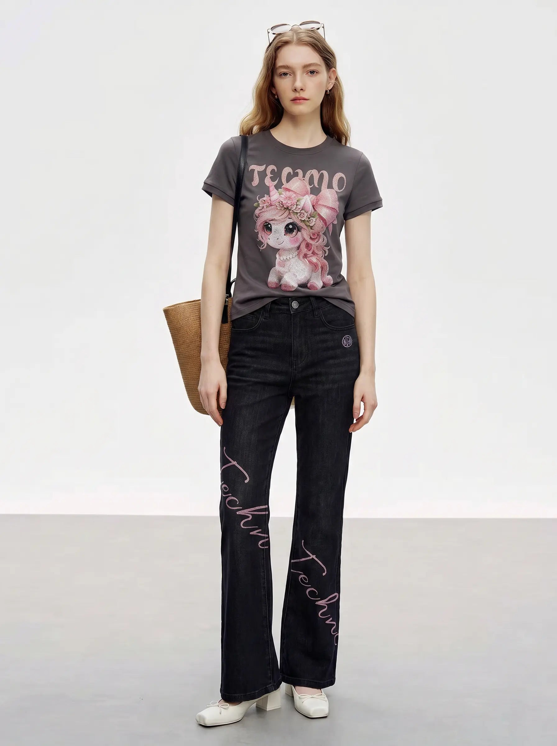 A model stands in a studio wearing a dark gray graphic t-shirt with a pink cartoon dog and the word 'TECNO', paired with black wide-leg jeans featuring pink script embroidery down the front, white shoes, and carrying a tan shoulder bag. TCH Rhinestone Unicorn Graphic Charcoal T-Shirt