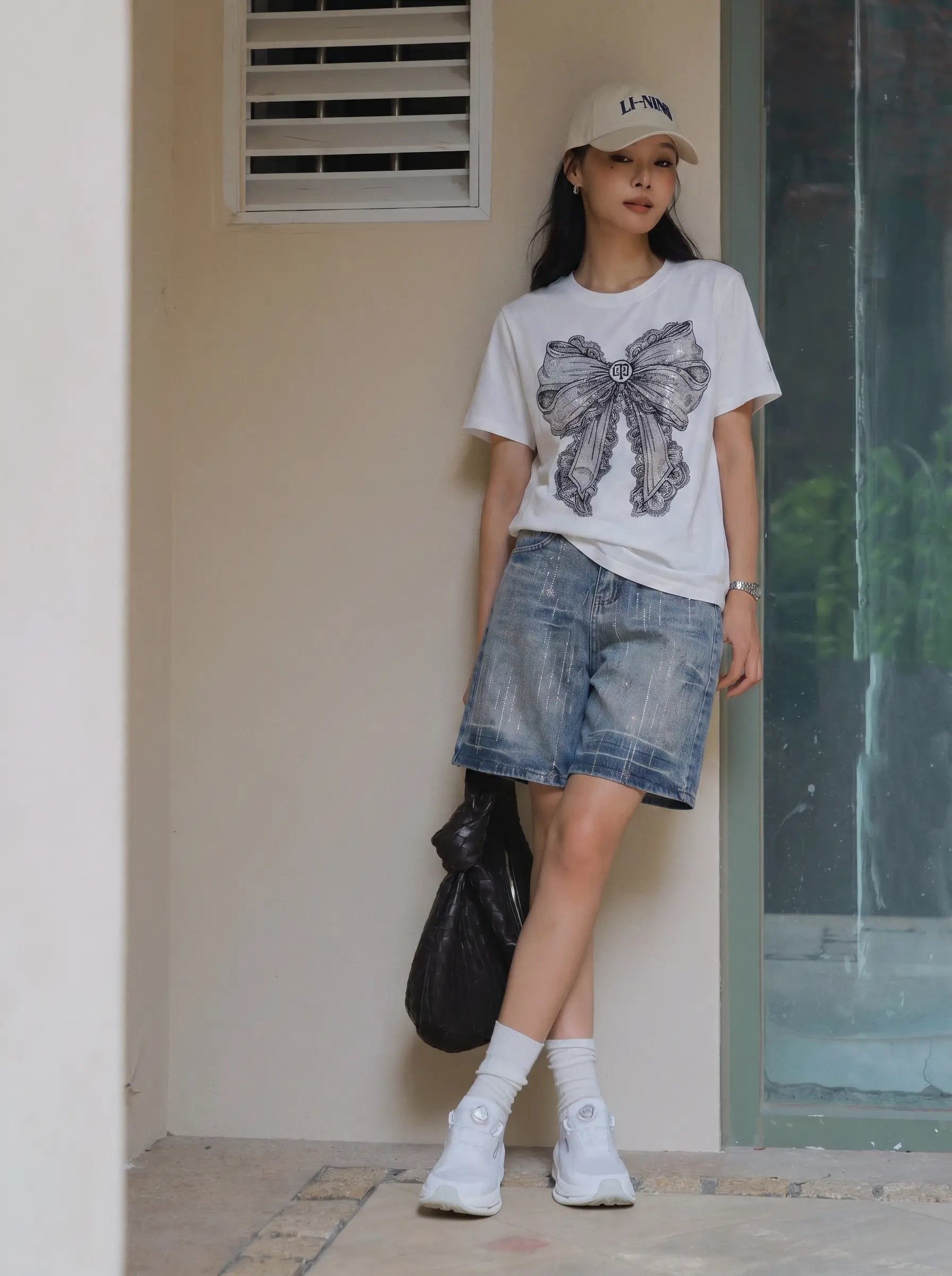 A woman stands in a doorway wearing a white graphic T-shirt with a large bow print, blue denim shorts, white socks, white sneakers, and a beige baseball cap. TCH Rhinestone Vintage Bow Graphic White T-Shirt